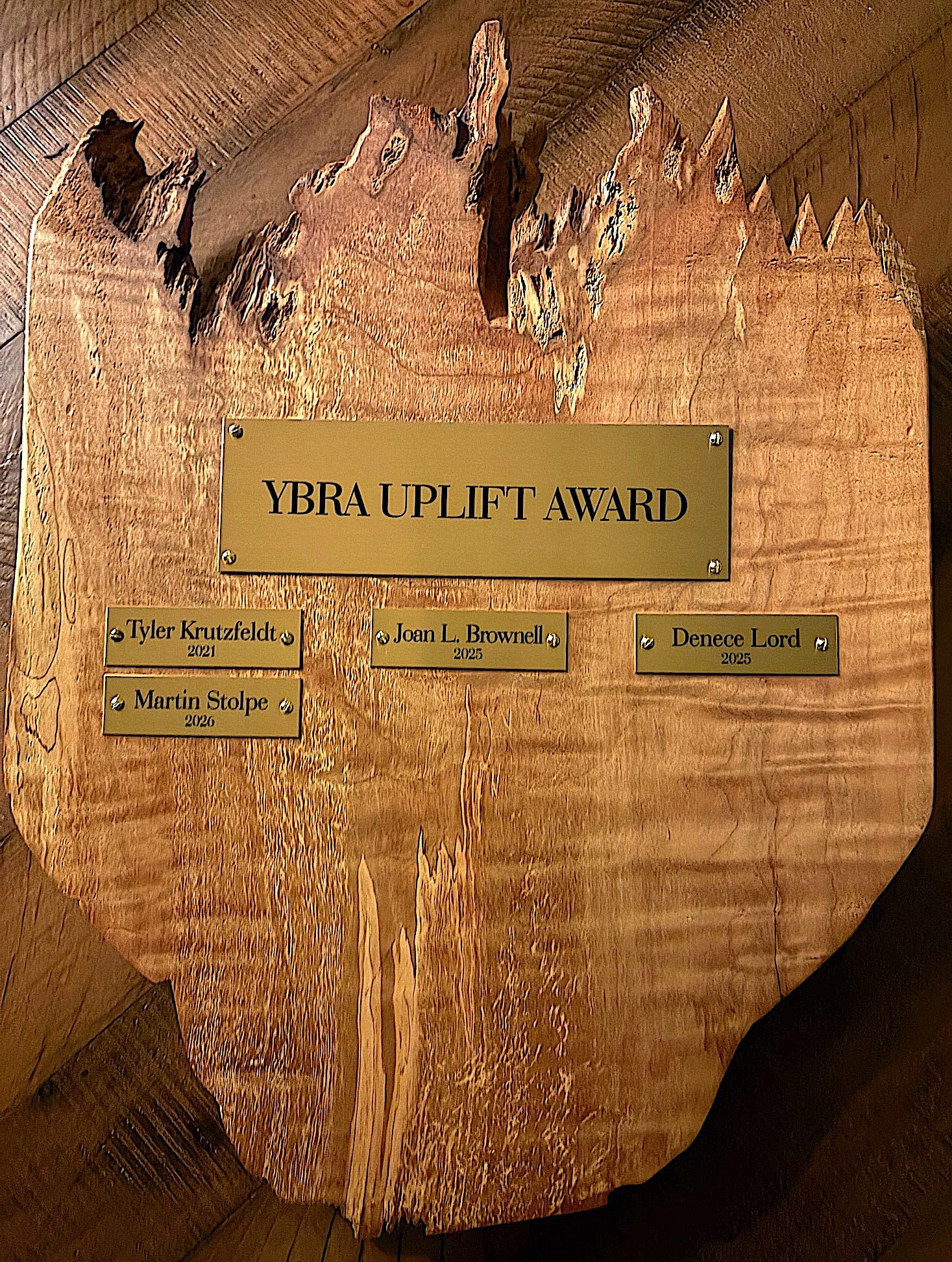 A wooden award plaque for the YBRA Uplift Award mounted on a slice of wood, with engraved names and years of recipients, including Tyler Krutzfeldt, Joan L. Brownell, Denece Lord, Martin Stolpe.