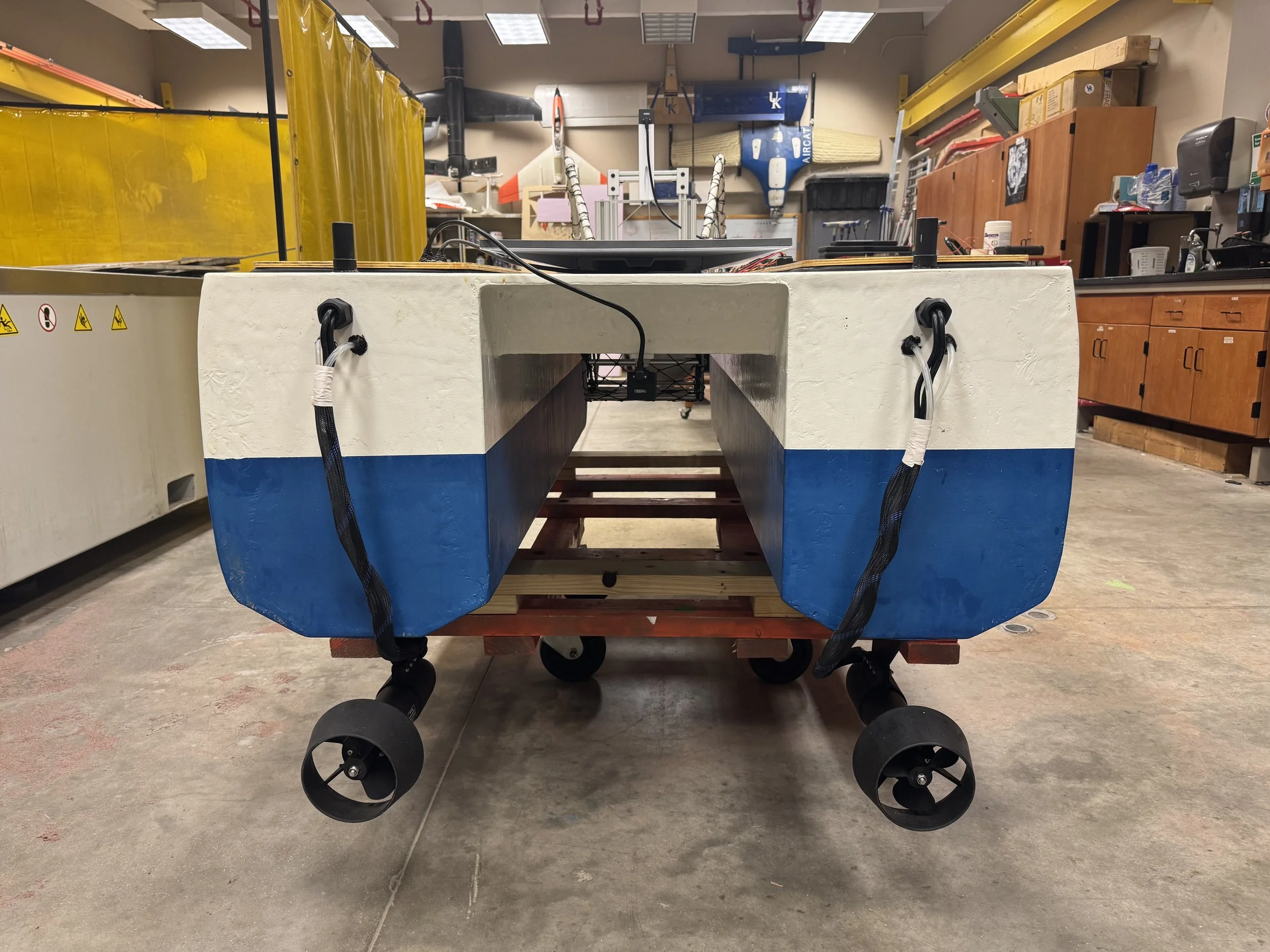 A large U-shaped boat hull with a white top and blue bottom, mounted on a wheeled cart in a workshop. The hull has black cables attached to the front and is surrounded by tools and equipment.
