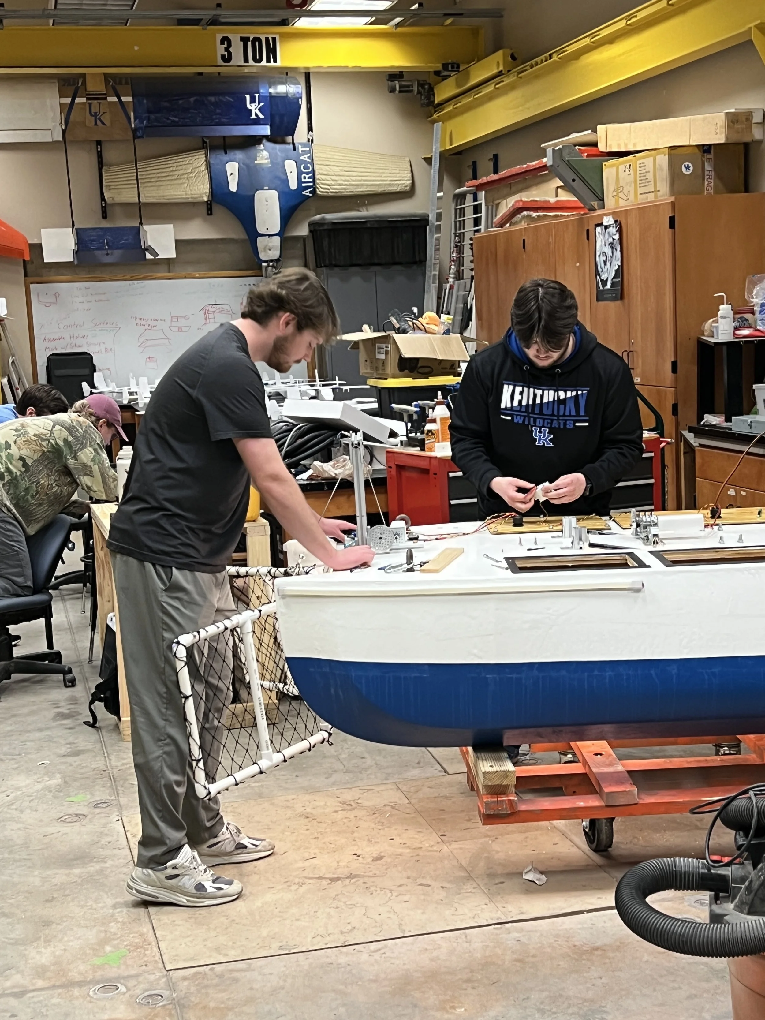 Two young men working on a boat in a cluttered workshop, with tools and equipment around them, and a yellow overhead crane labeled "3 TON".