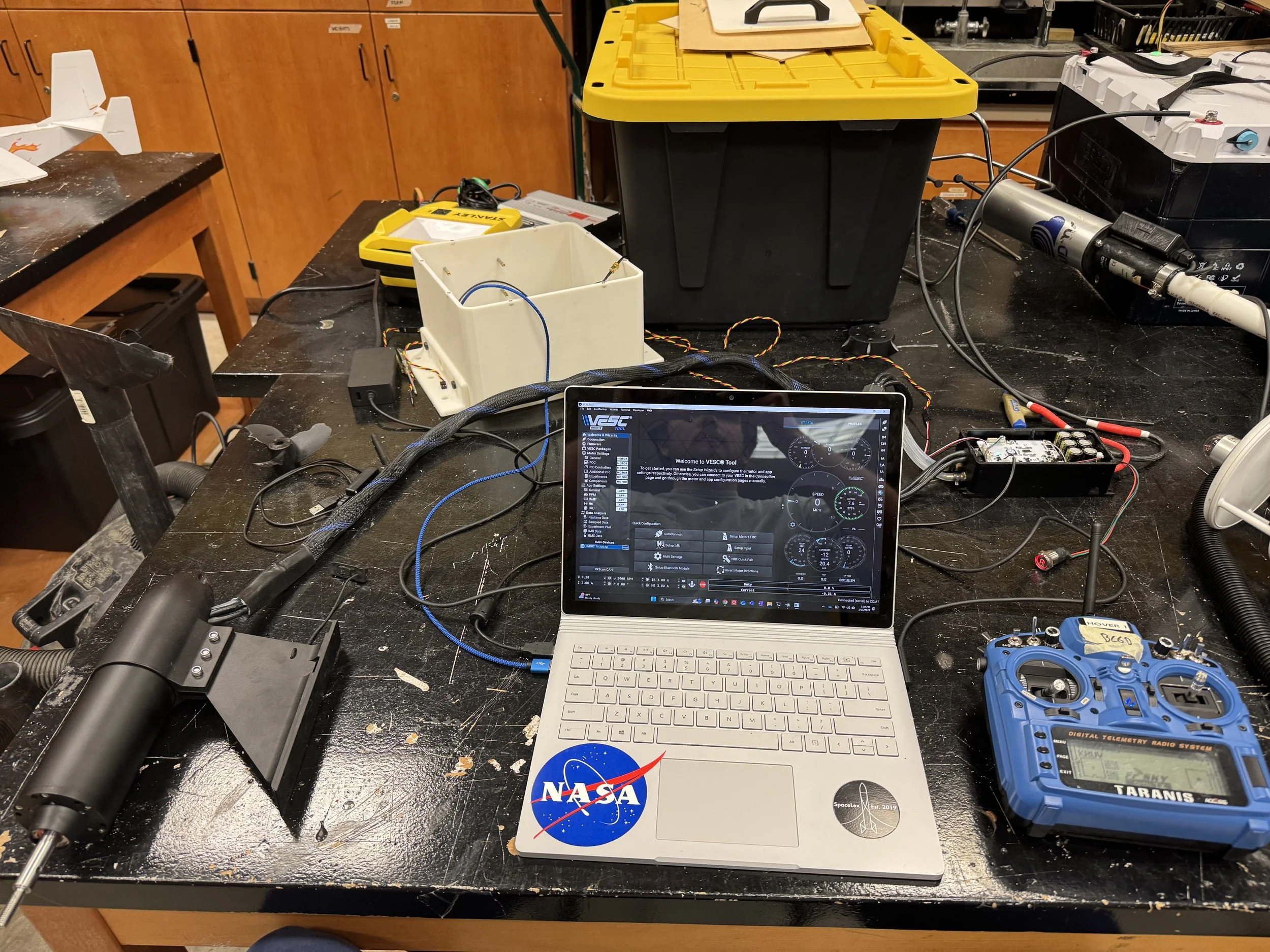 A workbench with various electronic and robotic equipment, including a laptop with a NASA sticker, a blue Taranis remote controller, a white box with wires, and other electronic components and tools.