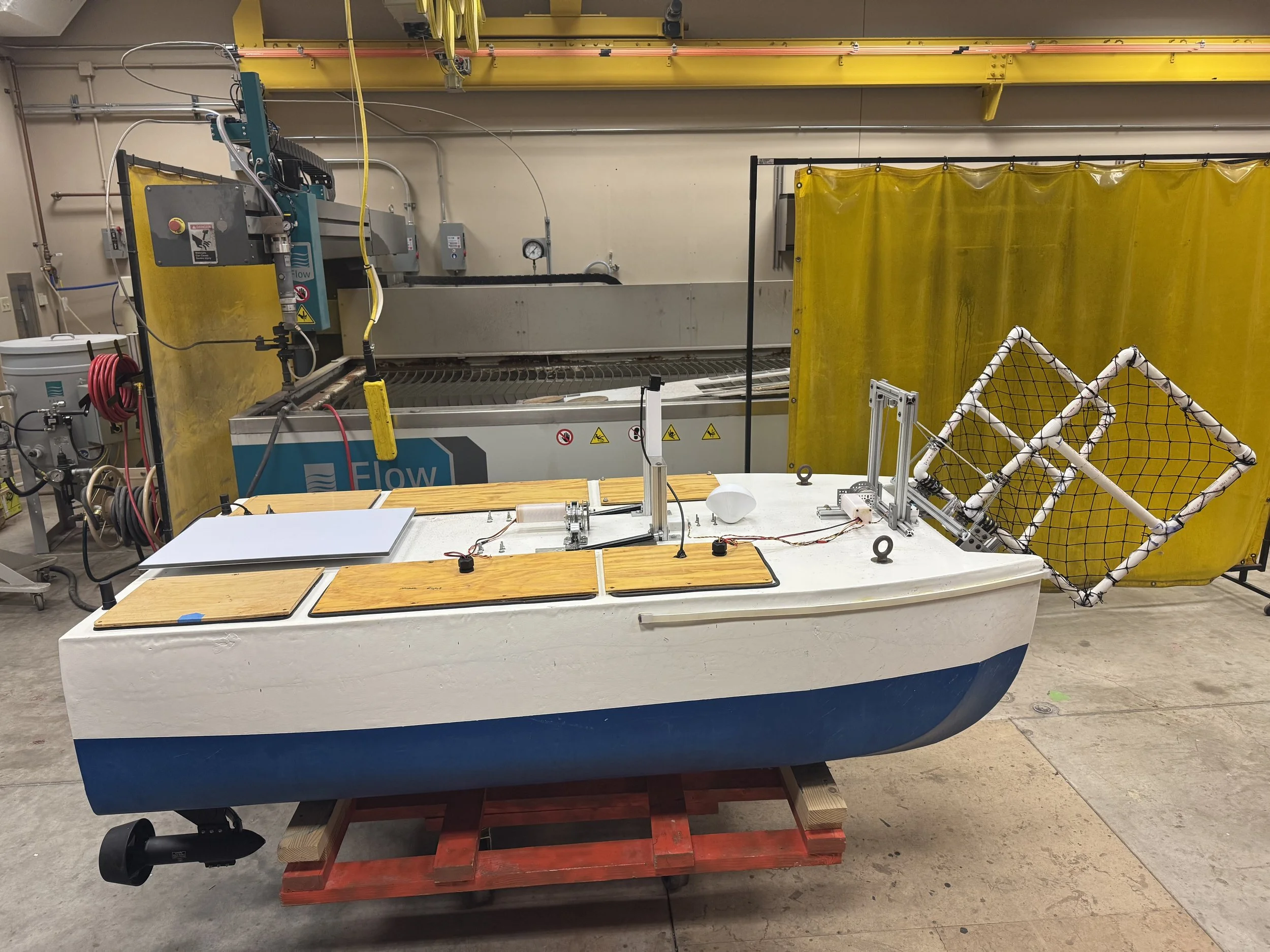 A small sailboat with a white hull and blue bottom, equipped with various robotic components, is set up indoors in a workshop or laboratory, with industrial equipment and yellow safety curtains in the background.