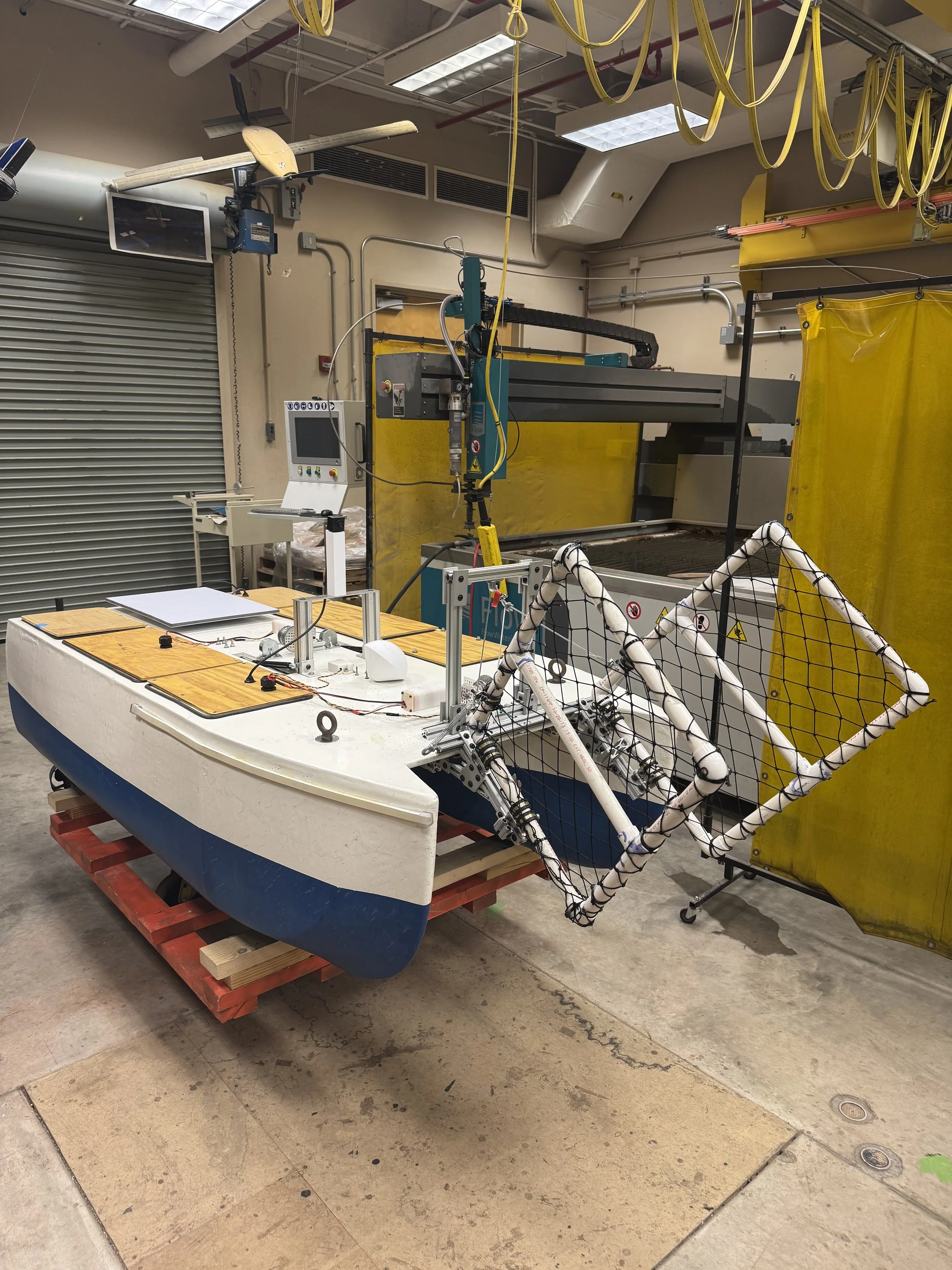 A boat fitted with a large robotic arm and equipment for testing or experiments, situated in an industrial lab with yellow safety barriers and overhead yellow cables.