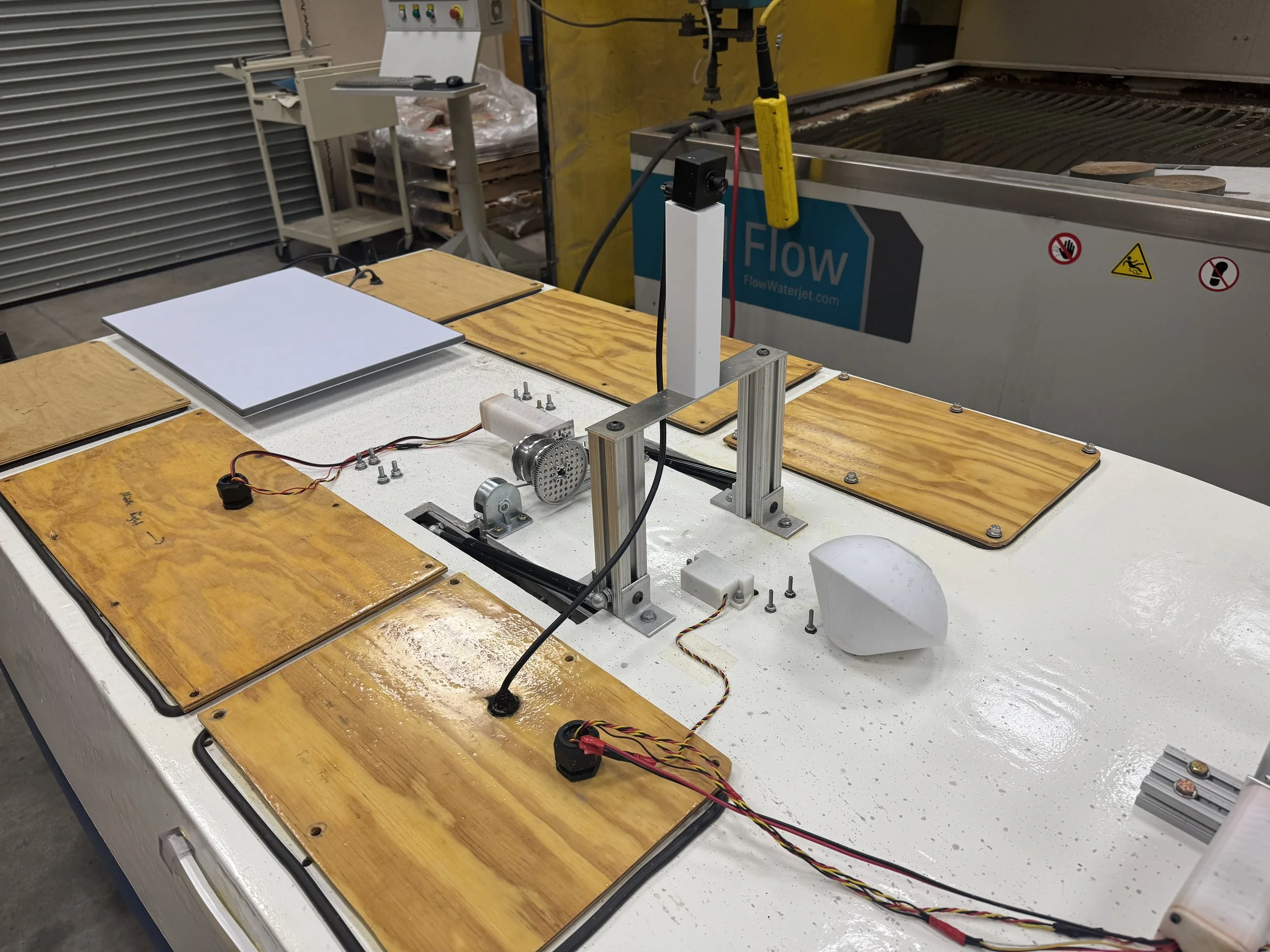 Workshop table with various electronic components, mounted on a white surface, including a stepper motor, wires, and a small metal frame, with a large industrial waterjet cutter in the background.