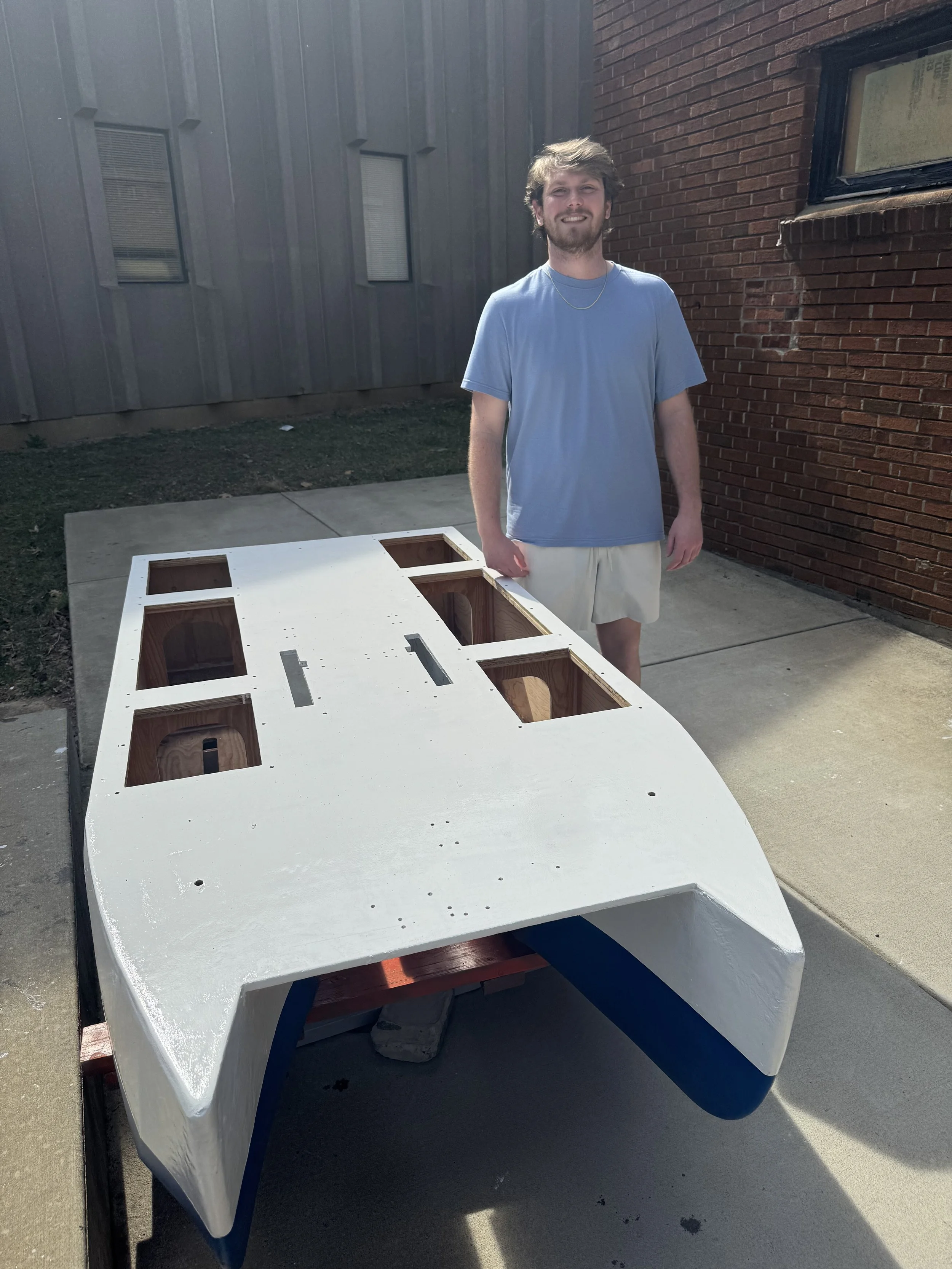 A young man with a beard and light brown hair standing outdoors on a concrete patio next to a partially assembled airplane fuselage with a white exterior, wooden interior, and blue underside. The man is wearing a light blue T-shirt, beige shorts, and