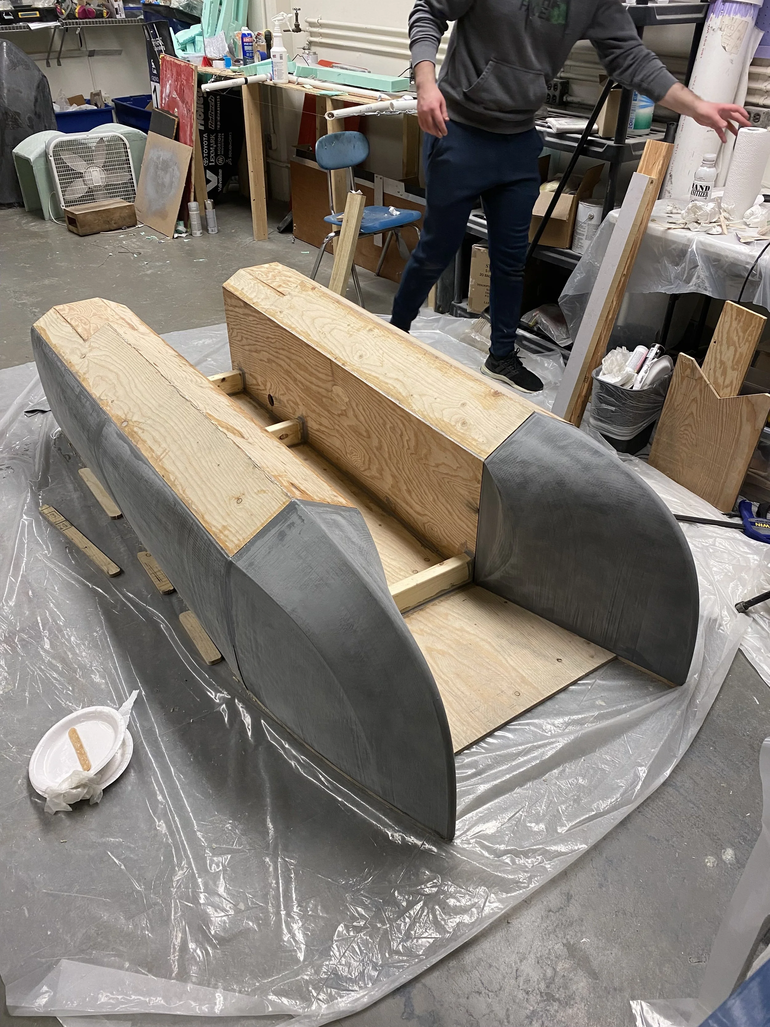 A woodworking project in progress with a wooden frame and curved metal sides on a plastic sheet. A person is working in the background in a workshop filled with tools, supplies, and furniture.