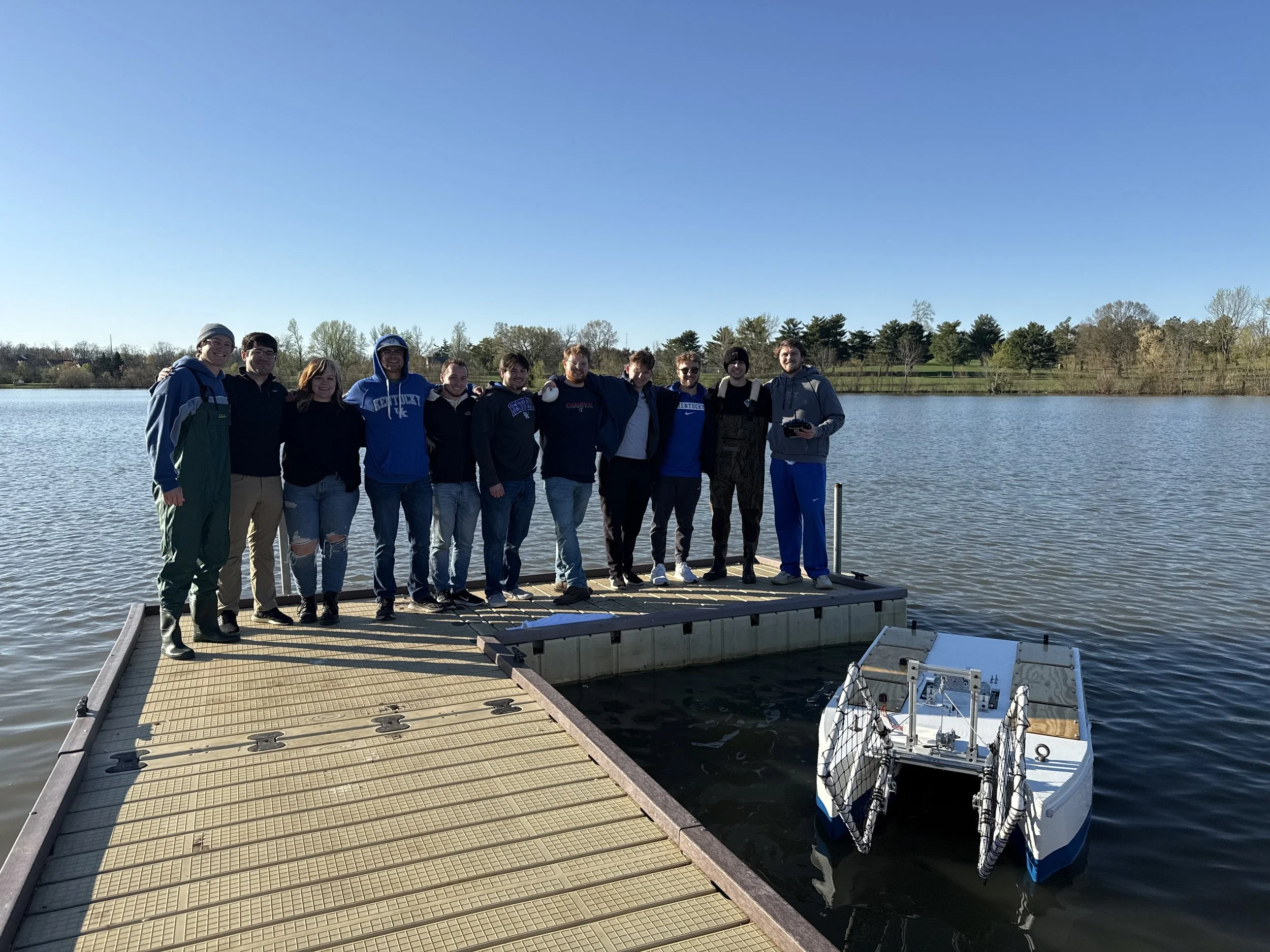 Group of people standing on a dock by a lake with a small autonomous boat nearby, during a sunny day.