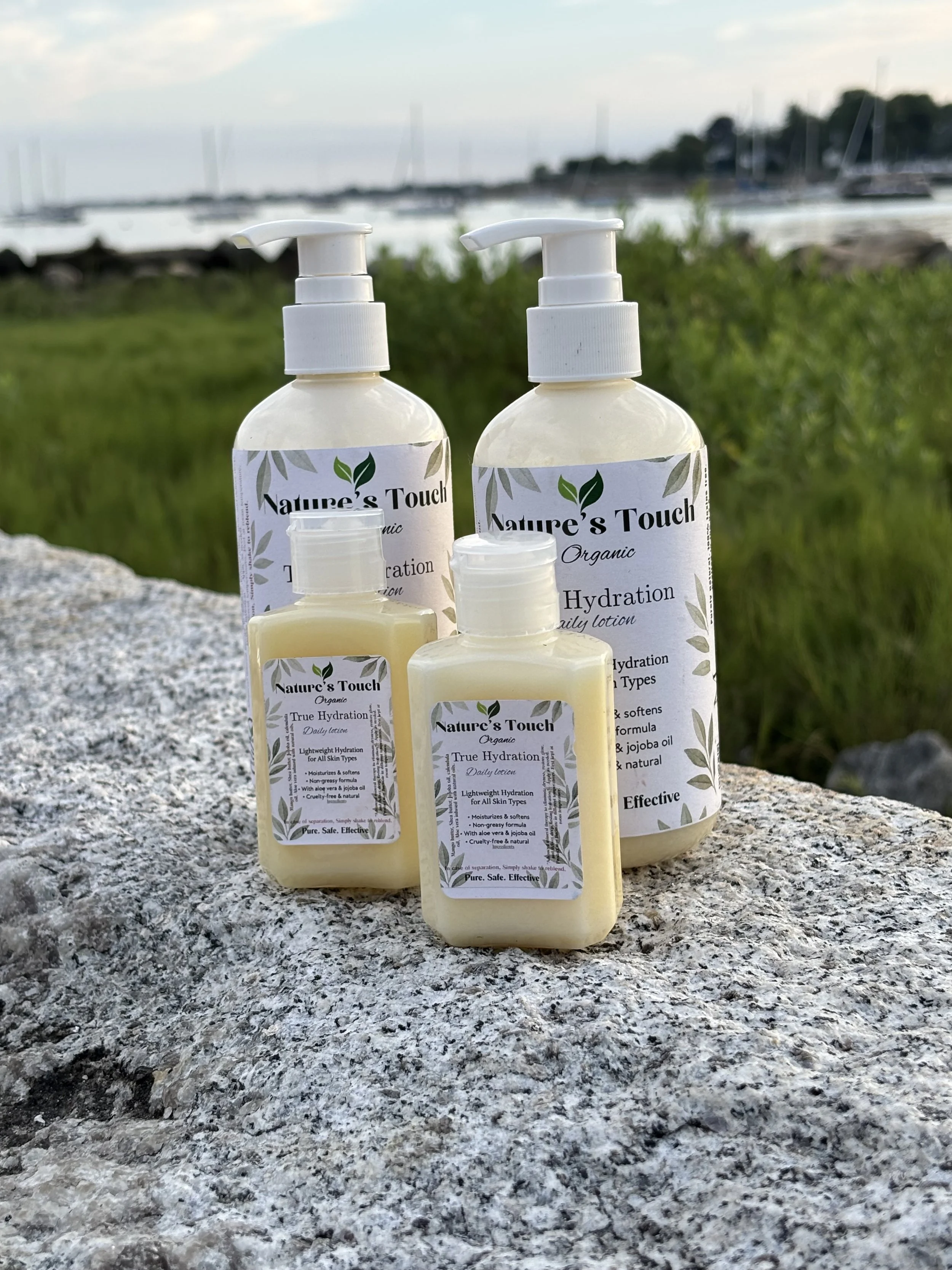 Four bottles of Nature's Touch organic hydration lotion placed on a granite surface outdoors, with a green natural landscape and marina in the background.