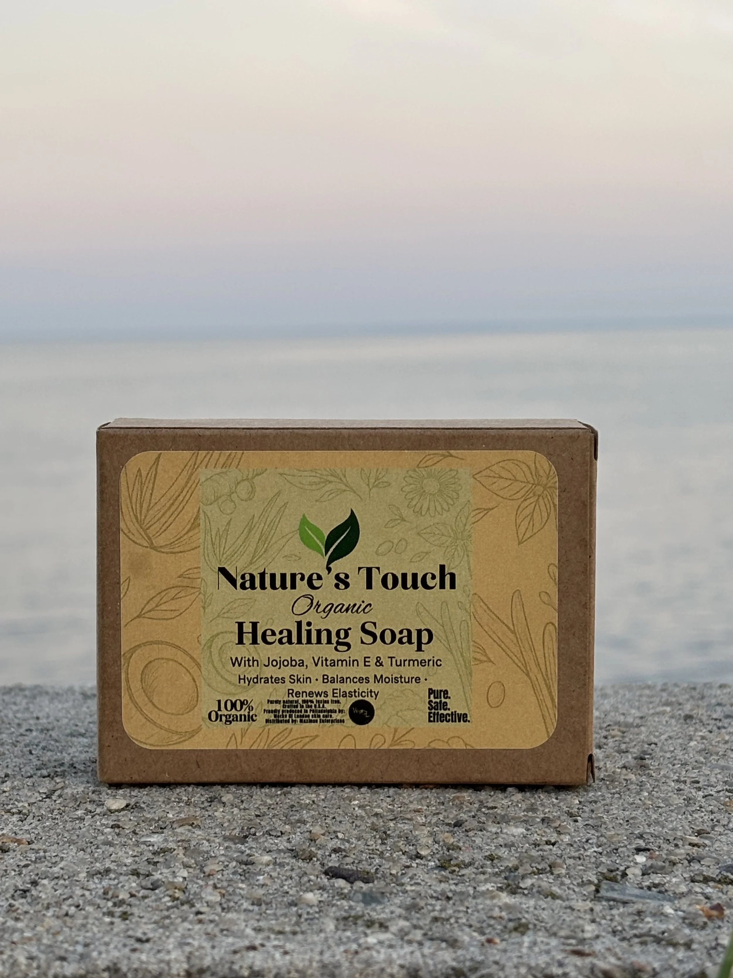 A box of Nature's Touch organic healing soap placed on a sandy surface with a blurred beach and ocean in the background.