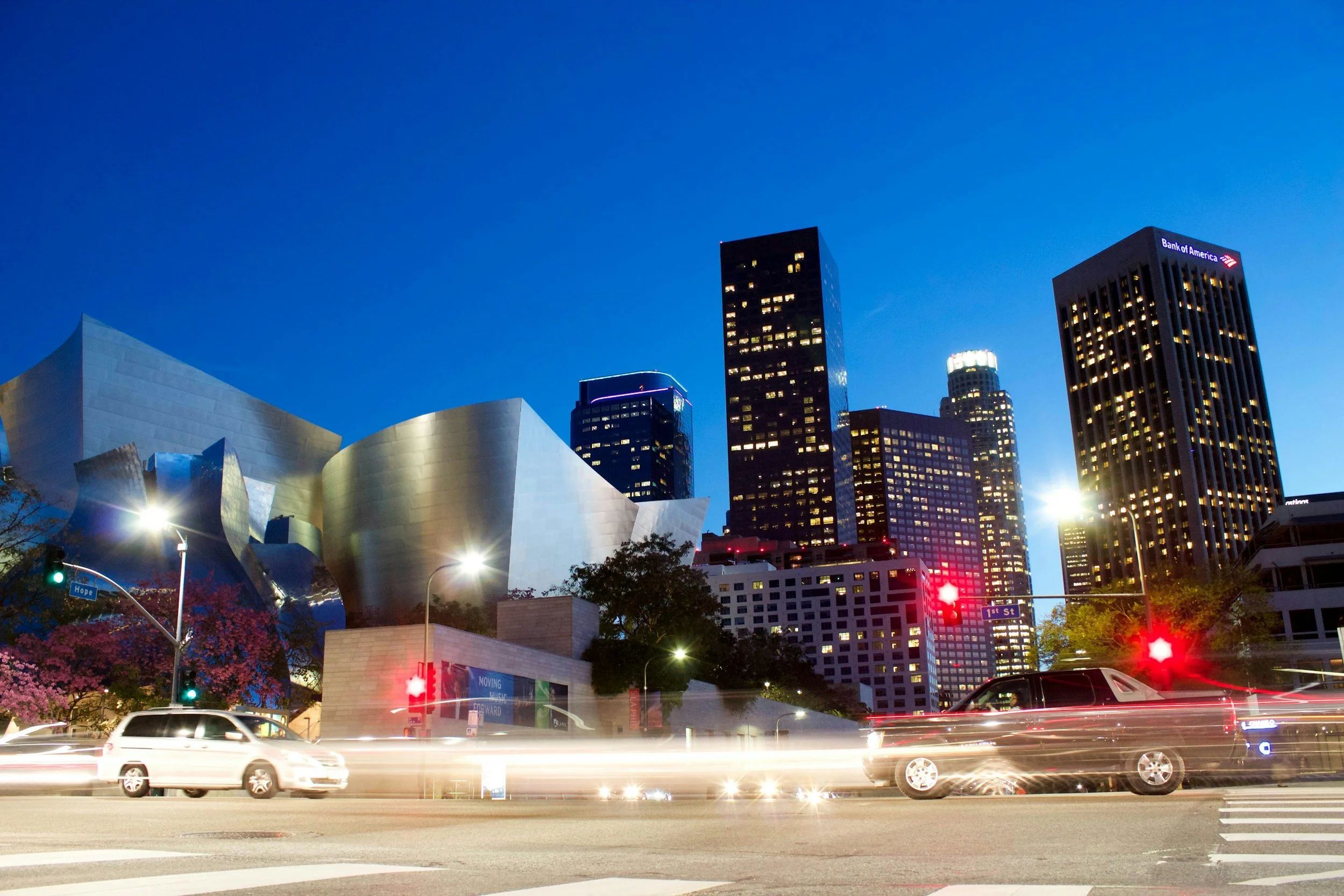 Nighttime cityscape with modern skyscrapers, including the Walt Disney Concert Hall, illuminated buildings, and streaks of car lights on the street.