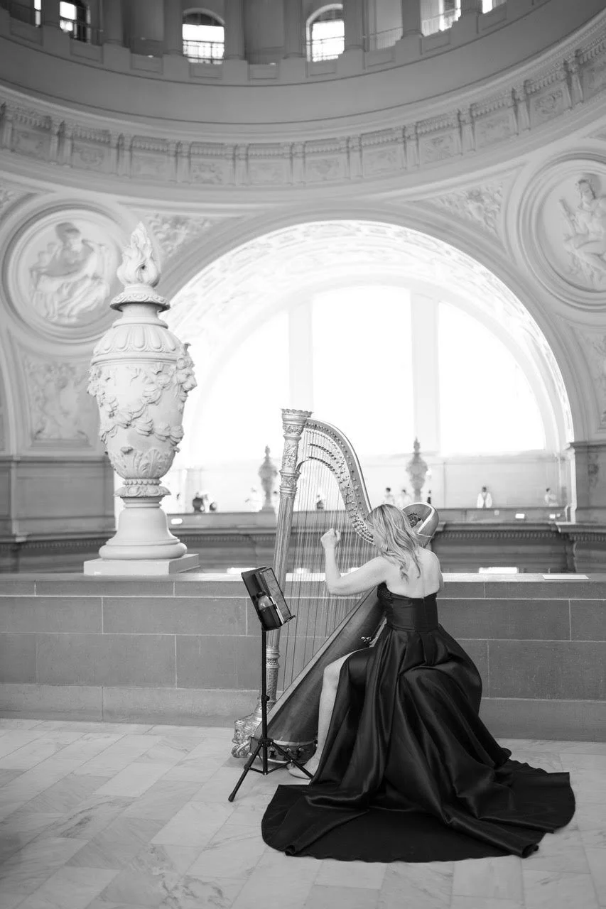 Black and white image of a harpist performing, wearing a black gown, inside of San Francisco City Hall, with large arches and plaster work behind her
