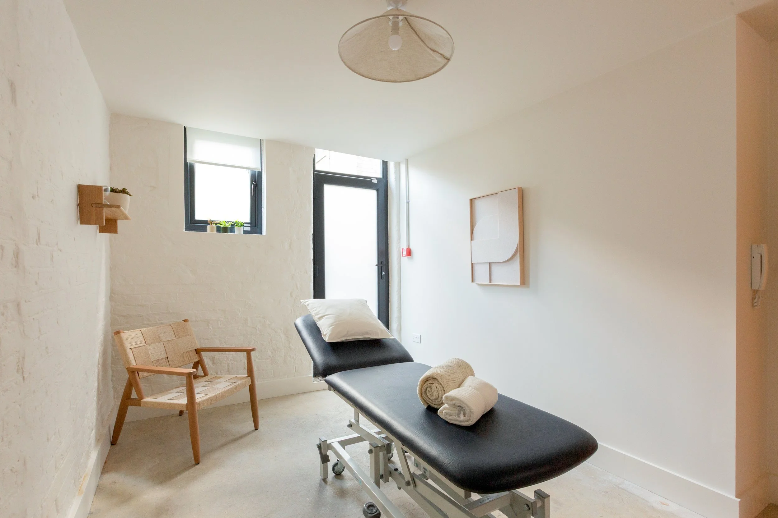 Minimalist therapy room with black massage table, rolled towels, and wooden chair, white walls, small window, and abstract art on the wall.