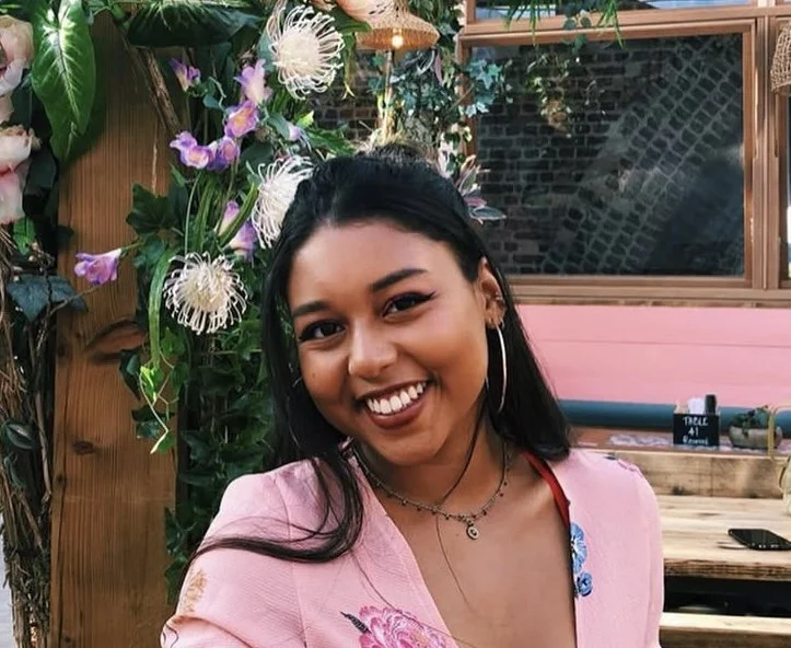 A young woman with long black hair, hoop earrings, and a pink embroidered jacket smiling at the camera. She is standing outdoors with a background of green plants and colorful flowers.