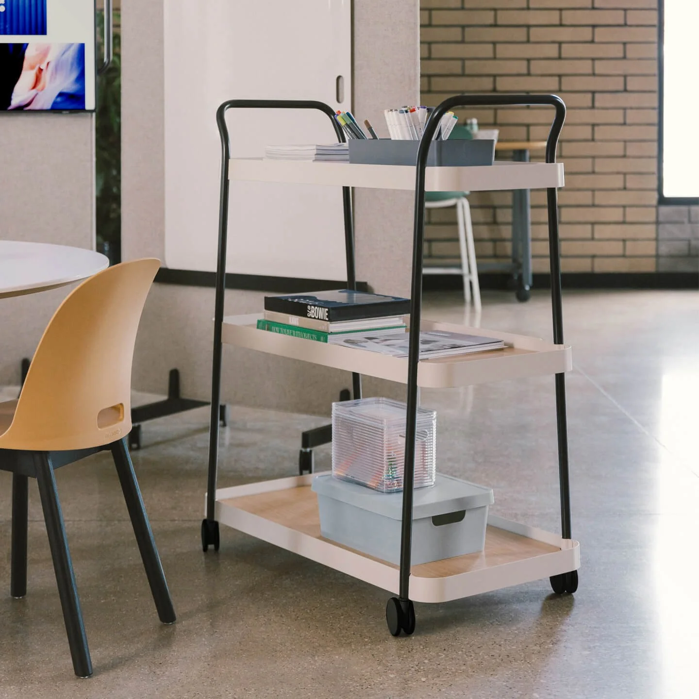 A multi-level cart holding books, magazines, and office supplies in an office space with a brick wall and window.