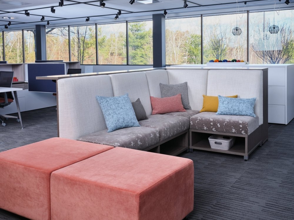 Modern office lounge with sectional sofa, colorful cushions, pink ottomans, and large windows showing trees outside.