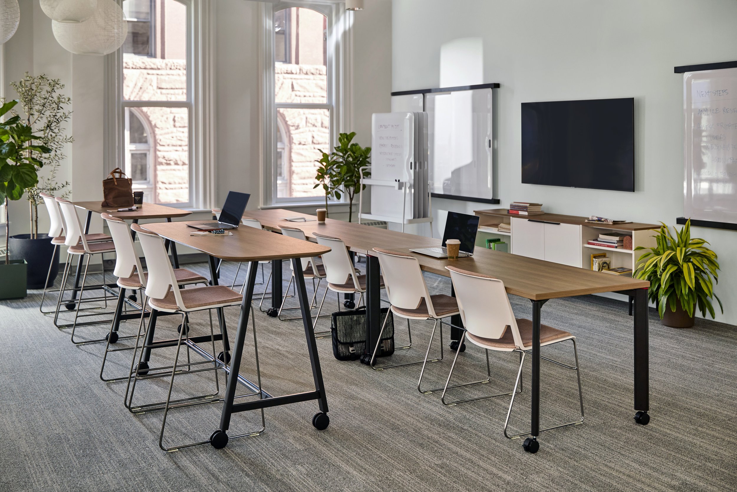 A modern, well-lit conference room with a long wooden table, white chairs, laptops, coffee cups, large windows with outdoor views, and whiteboards on the wall.