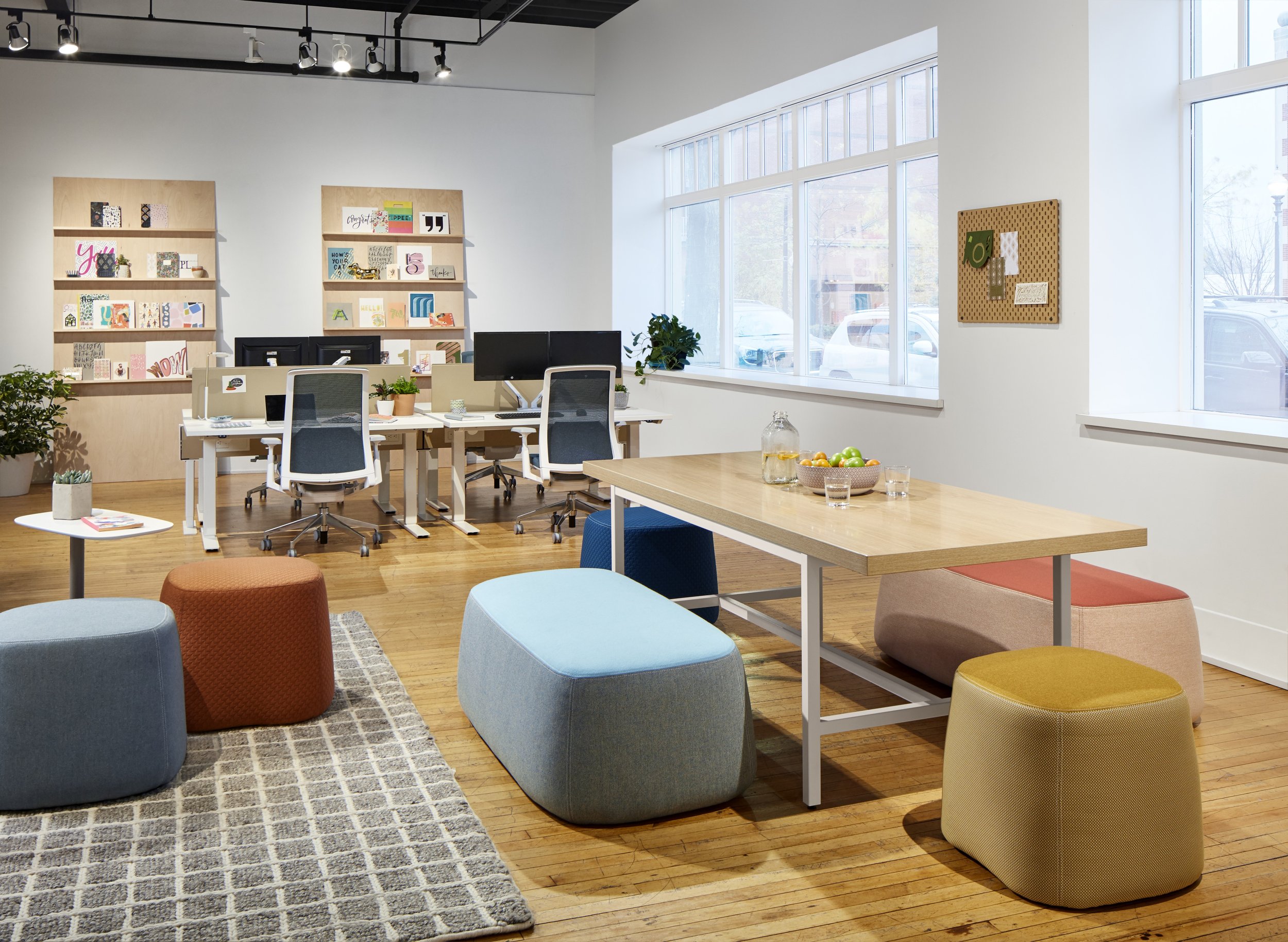 Modern office space with white walls, wooden floors, multiple desks, office chairs, colorful seating poufs, shelves with decor, a wooden table with fruit, and large windows letting in natural light.