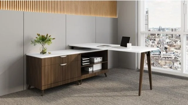 Modern office corner with a white L-shaped desk, a wooden cabinet with three drawers, a laptop, a white coffee mug, and a small plant on the desk. Large window with city view in the background.