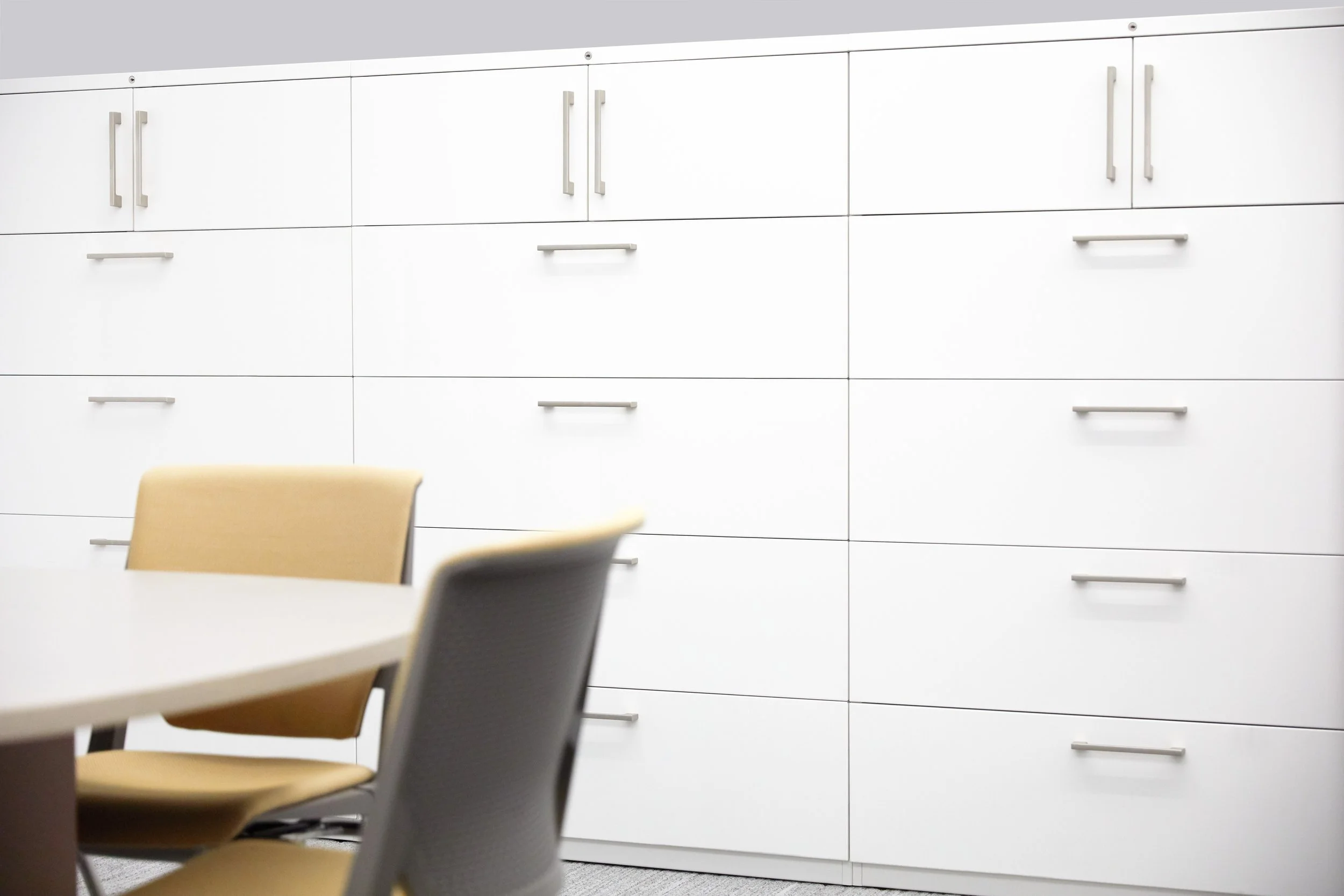 Empty office or conference room with white cabinets or lockers on the wall and beige chairs around a table.