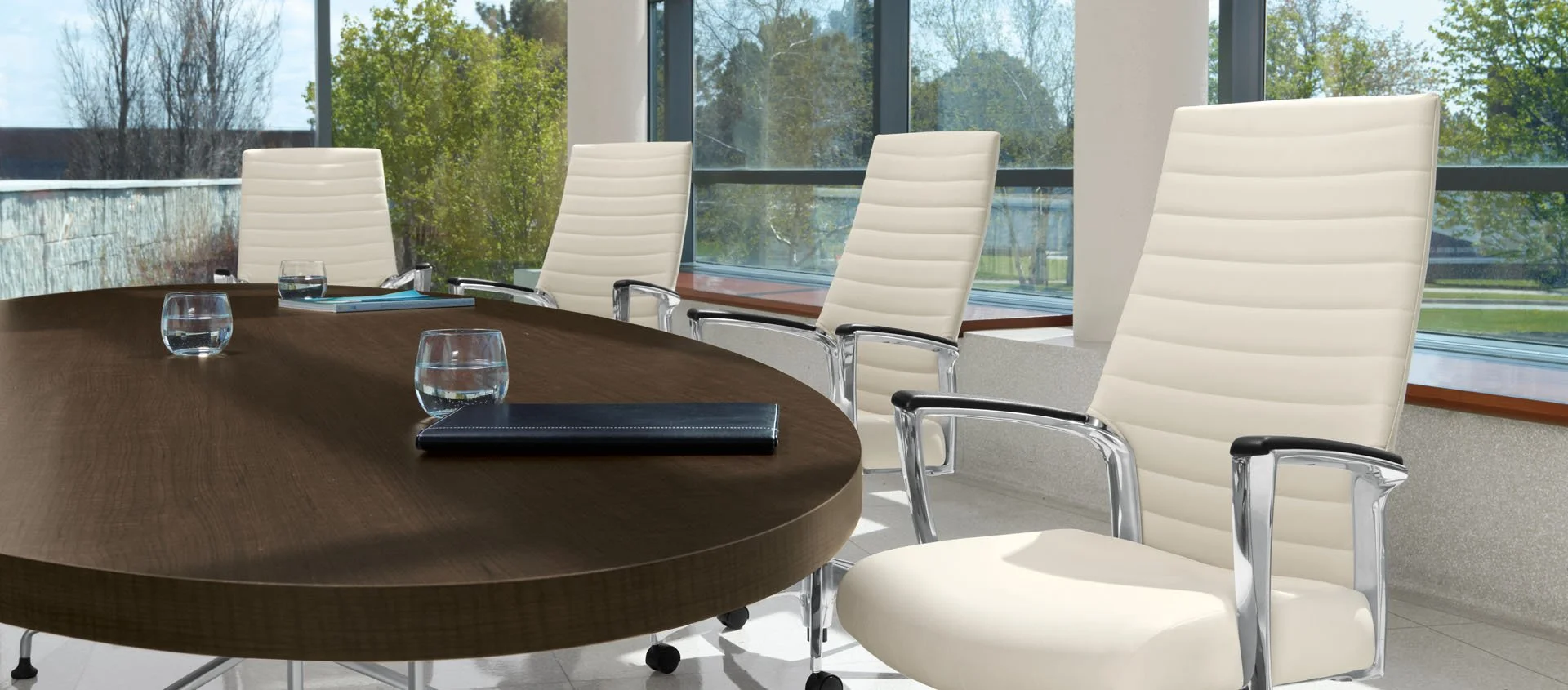 Conference room with large wooden table, four white leather chairs with chrome armrests and a glass of water on the table, large windows showing trees and a sunny day.