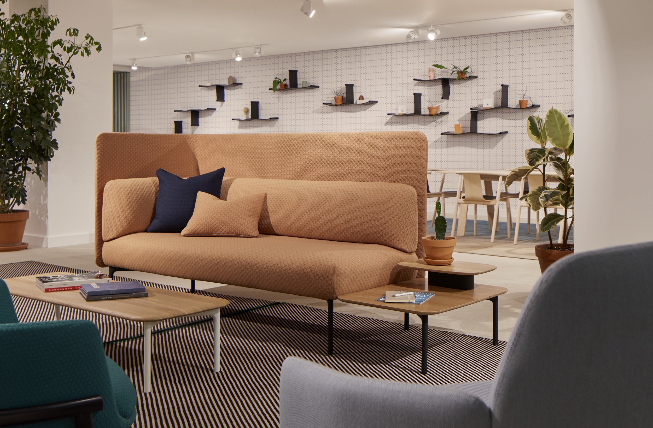 Modern living room with beige couch, blue and beige throw pillows, potted plants, and wooden coffee tables. In the background, a dining area features a table and chairs, with decorative shelves on the wall.