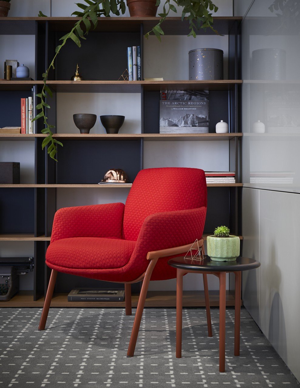 A modern room featuring a vibrant red armchair with wooden legs, a small black side table with a green potted succulent, a geometric decorative object, and a background of a wooden bookcase filled with books, vases, and decorative objects, with a gra