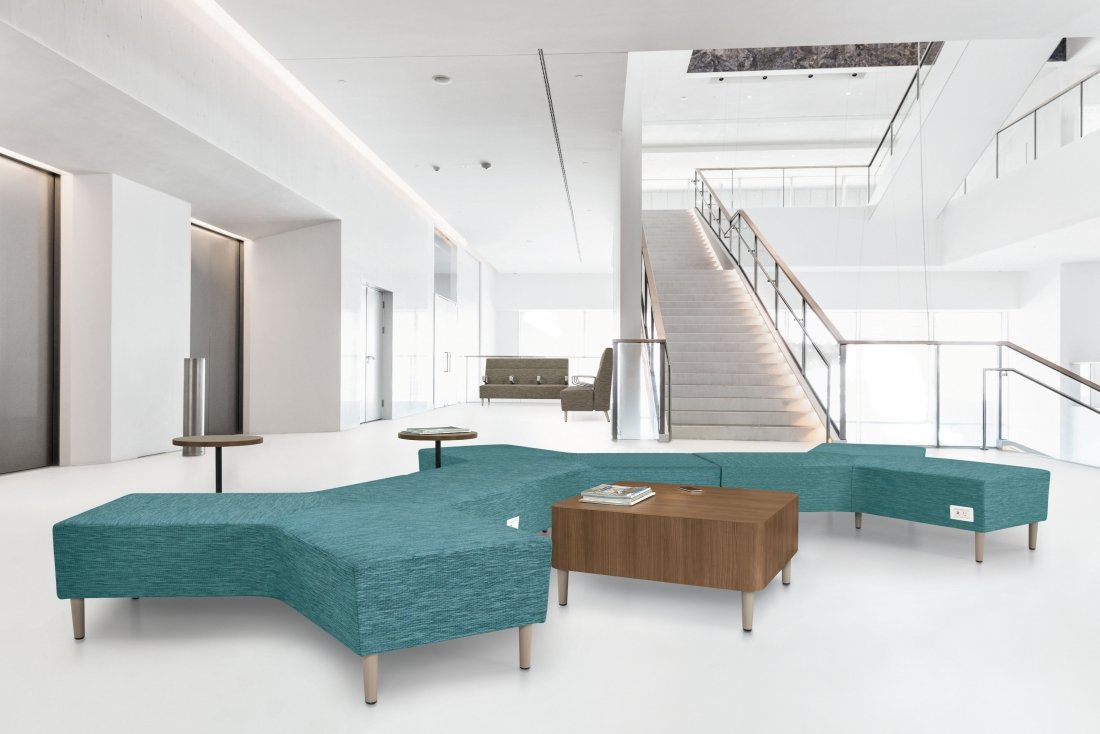 Modern, minimalist hotel lobby with turquoise sectional sofa, wooden coffee table, gray chairs, elevator doors, and staircase with glass railings in bright, open space.