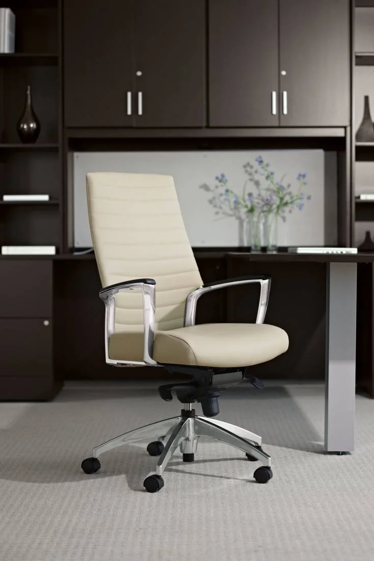Cream-colored office chair with leather upholstery and metal armrests in front of a dark wood office desk and cabinets, with a wall-mounted monitor and decorative vases.