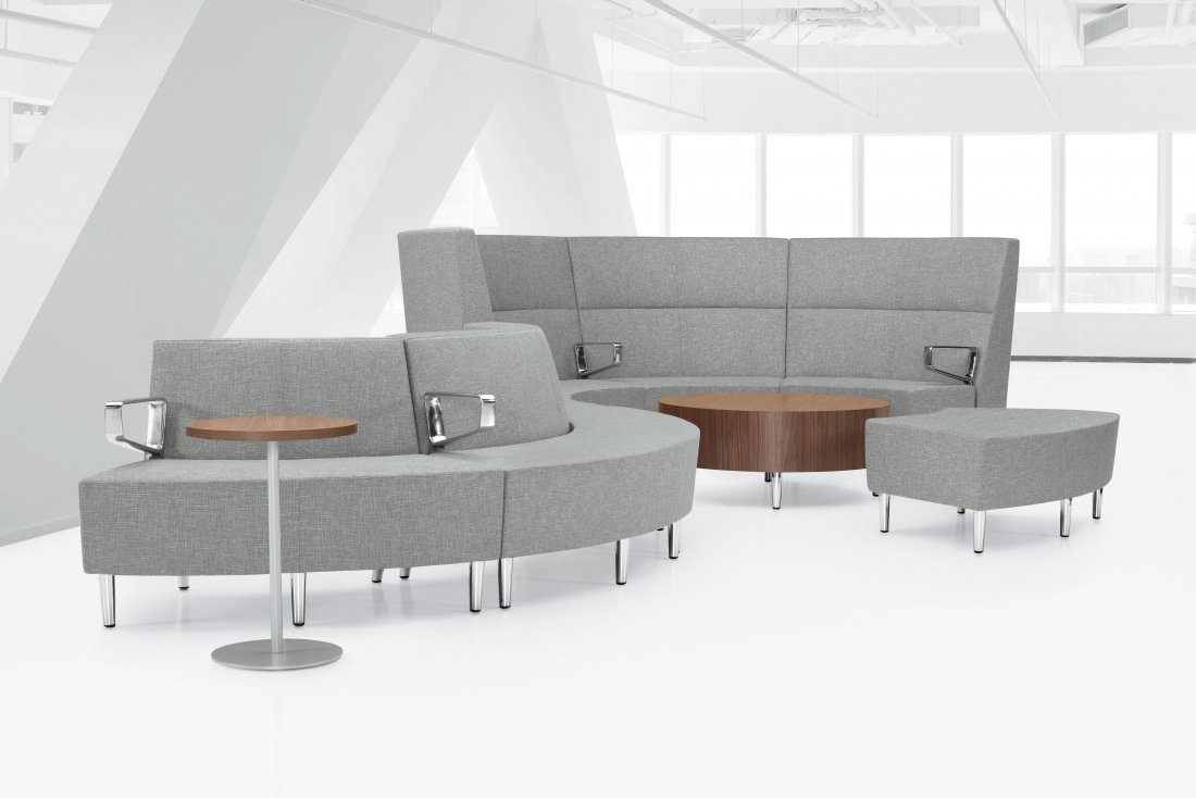 Modern office lounge seating with gray curved sofas, a small round wooden table, an ottoman, and a side table in a bright office space with large windows.