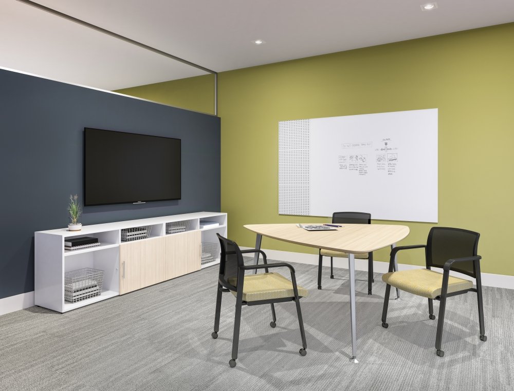 A small meeting room with a round table, three chairs, a whiteboard on a green wall, a large flat-screen TV on a dark blue wall, and a white cabinet with storage drawers.