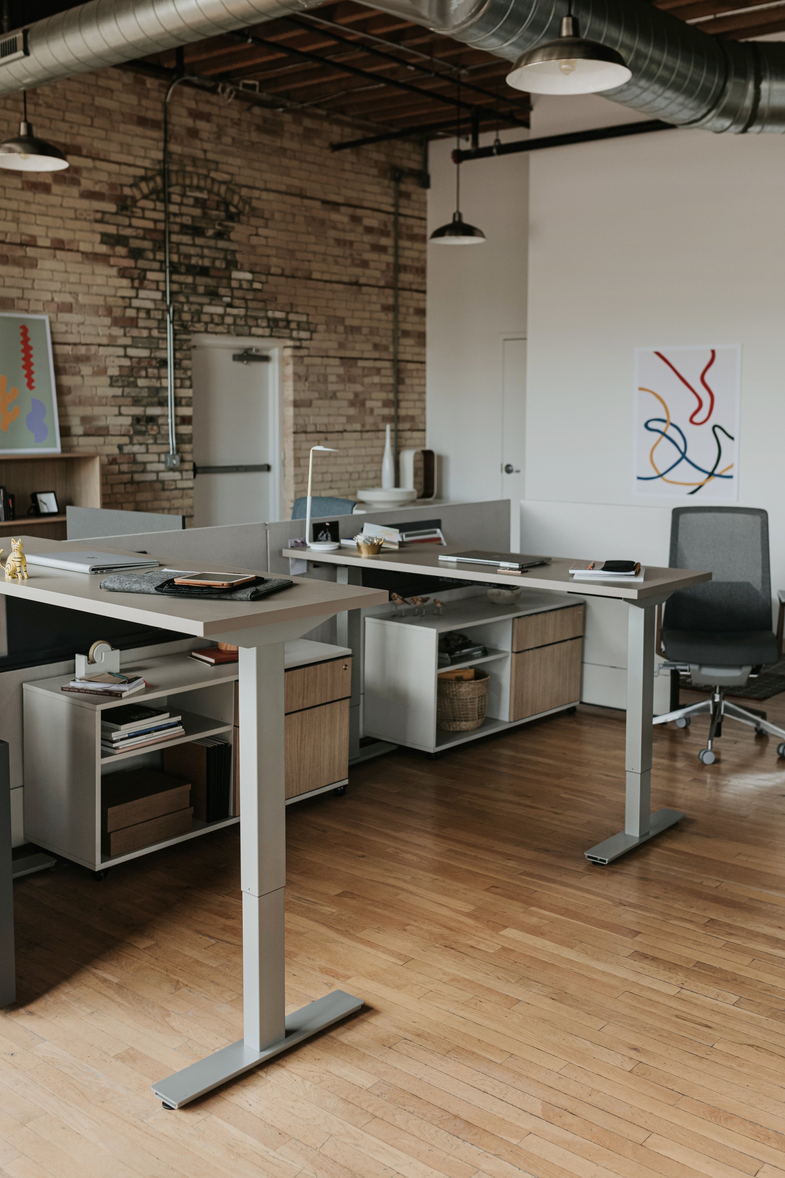 Modern office with three adjustable desks, a black office chair, brick wall, and colorful abstract art.