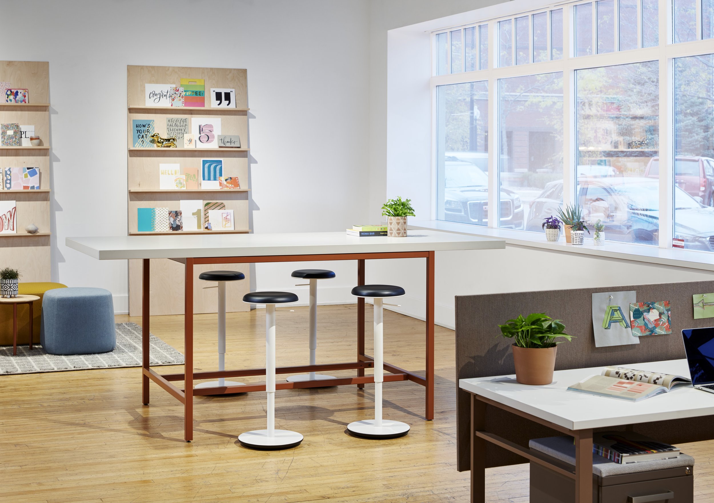 Modern office space with a high white table, three black and white bar stools, a seating area with a yellow ottoman and a blue pouf, a window ledge with potted plants, artwork on the walls, and a laptop on a desk.