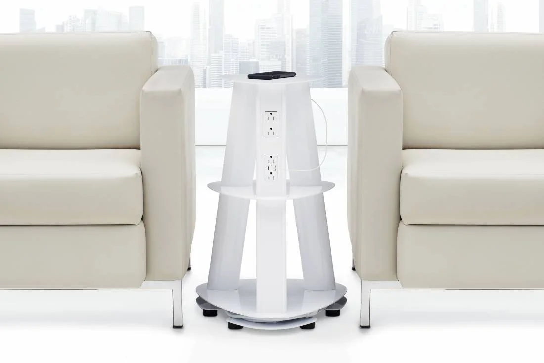 Two white armchairs with a round white side table between them, holding a smartphone, an outlet strip, and a cable, in front of a large window with a city skyline in the background.