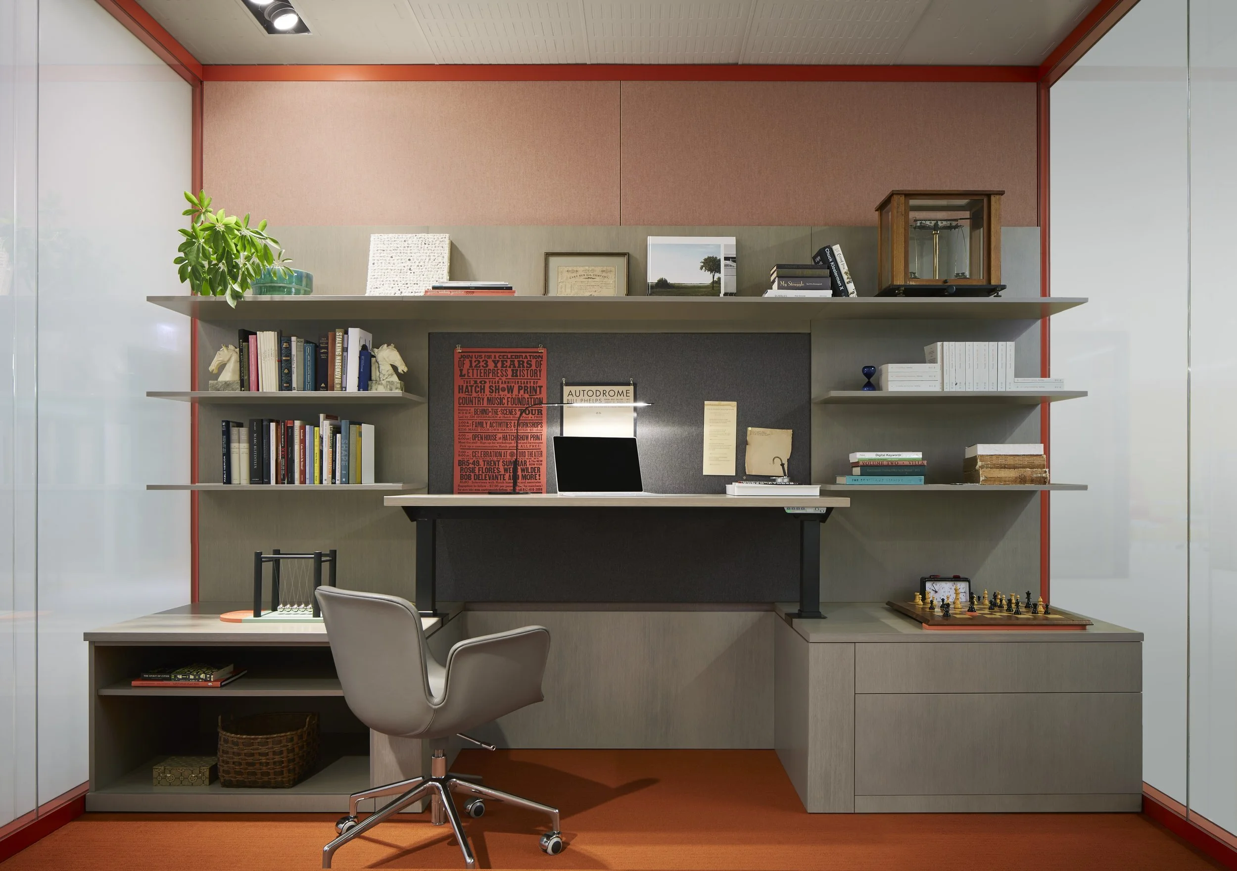 Modern office workspace with a gray desk, gray chair, and multiple shelves filled with books, framed pictures, and decorative items. There is a chess set on the right side of the desk and an open laptop in the center.