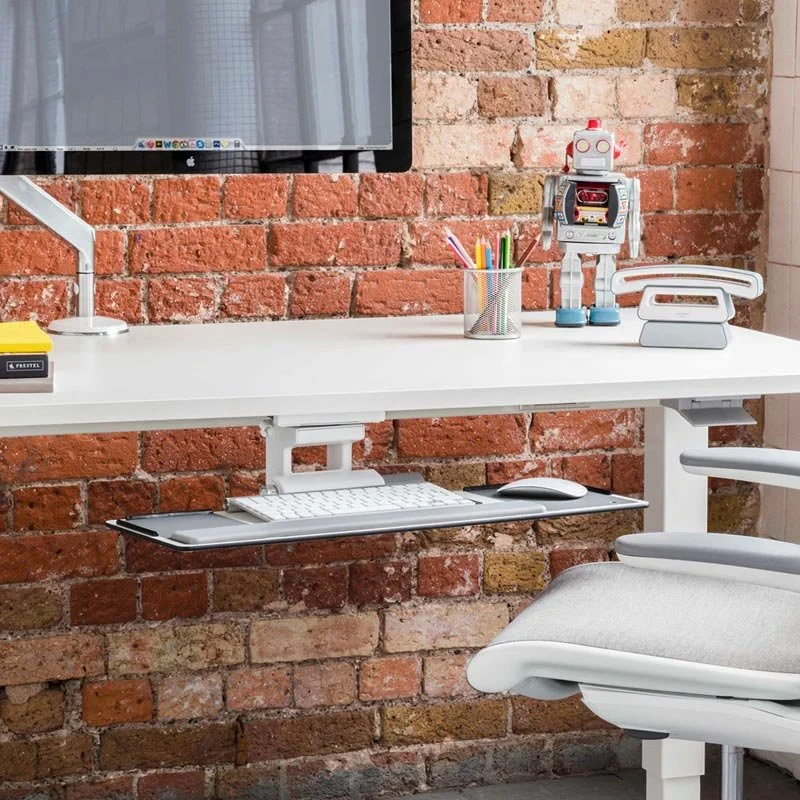 Modern office desk with a computer monitor, colorful pens in a metal holder, a robot figurine, a white keyboard on a sliding shelf, and a white office chair with a brick wall in the background.