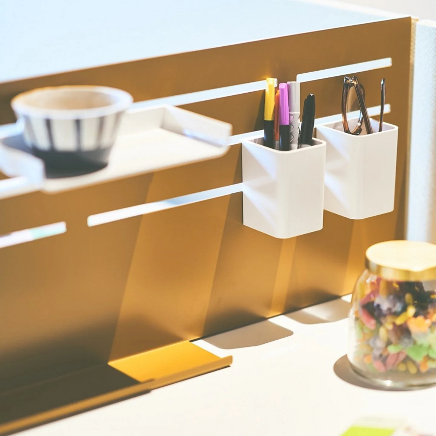 A desk organizer with compartments holding pens, markers, and reading glasses, with a small bowl and a jar of colorful candies nearby.