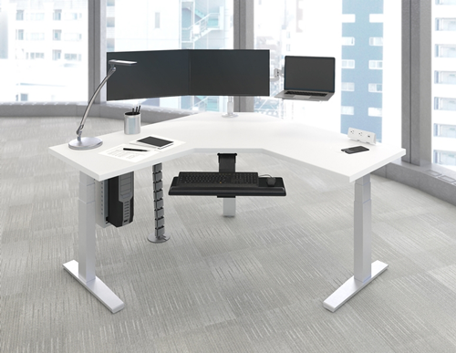 Modern office desk with dual monitors, a desk lamp, keyboard, mouse, and office supplies, set in a bright office with large windows.