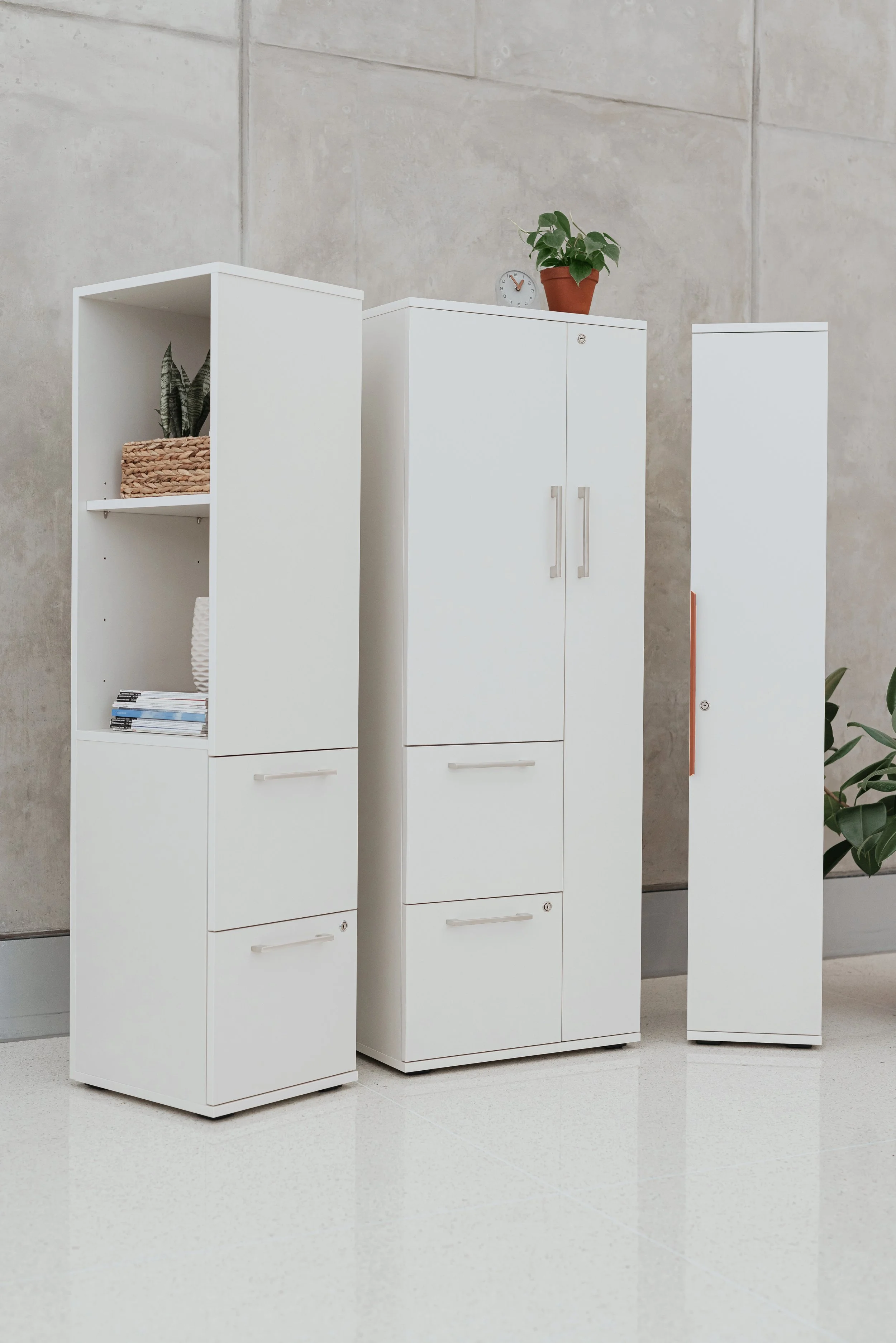 Three white storage cabinets with plants and a clock on top, set against a beige tiled wall.