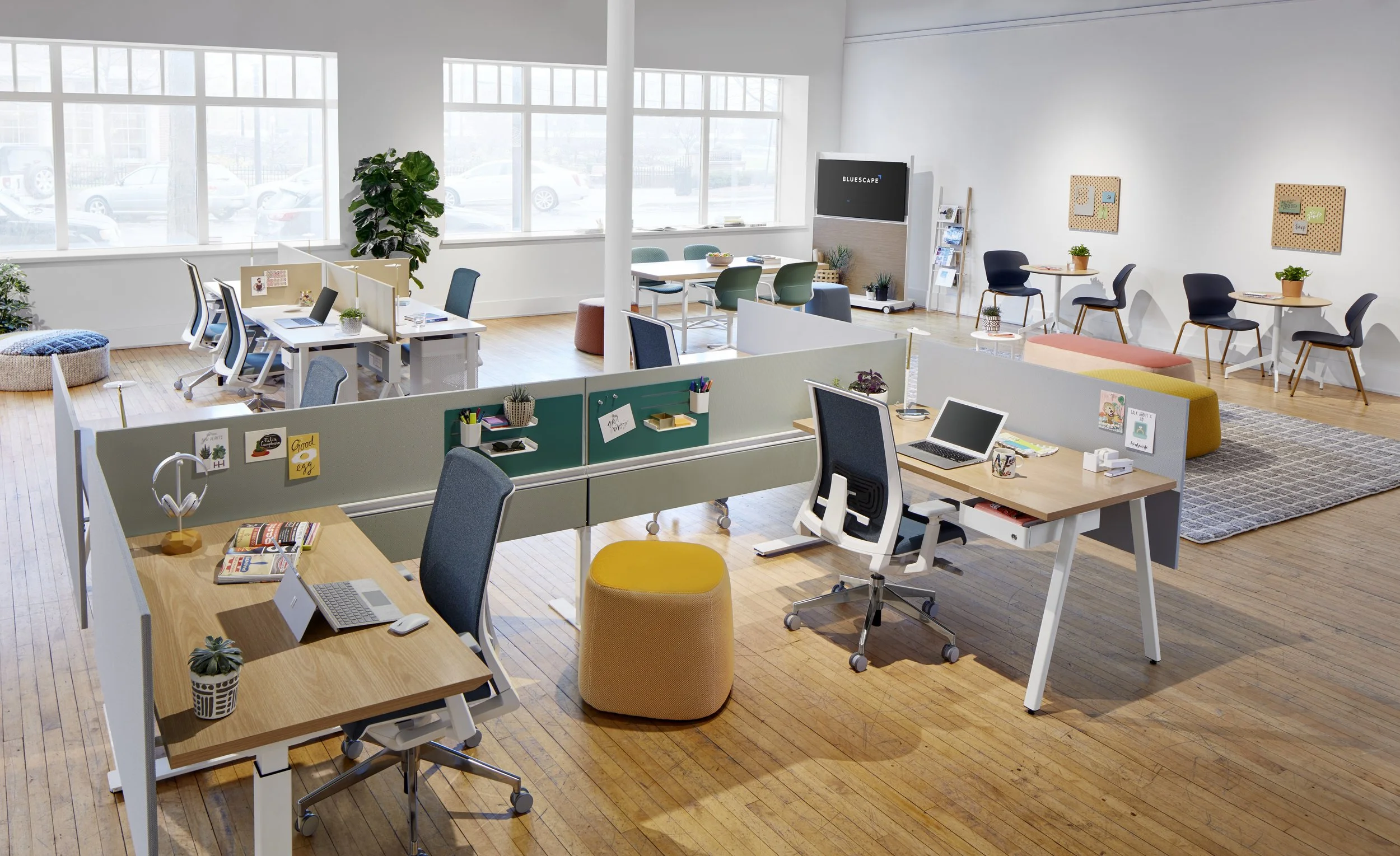 Open office with multiple workstations, desks, chairs, and seating areas, large windows, plants, and decor