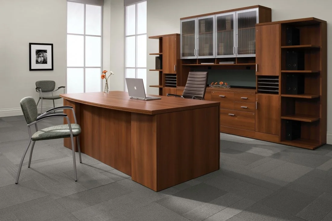 Office with wooden desk, gray office chair, two guest chairs, and large wooden cabinets with glass doors, in front of large window with frosted glass.