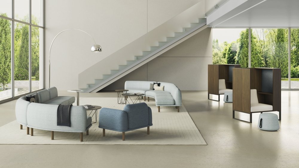 Modern living room with large windows, grey and blue sofas, a white rug, and a staircase with glass railing.