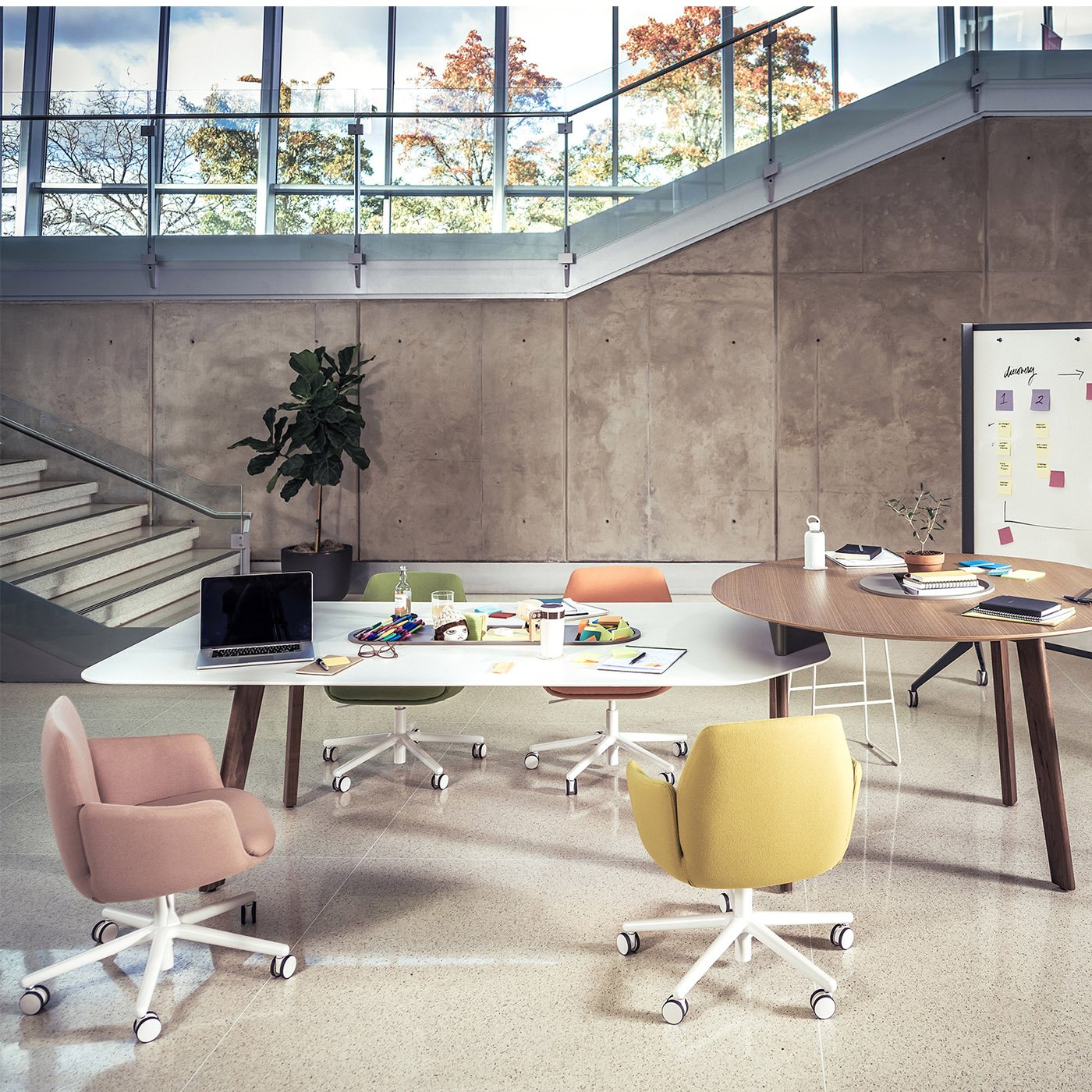 Modern office space with colorful chairs, a white and a wooden table, a laptop, stationery, notebooks, a whiteboard with sticky notes, and a large window showing trees outside.
