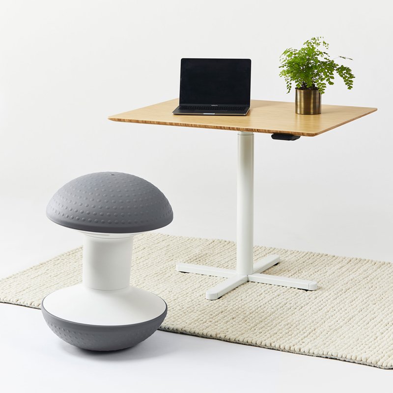 A minimalist workspace with a wooden table holding a black laptop and a potted green plant, and a white and gray stool on a cream rug.