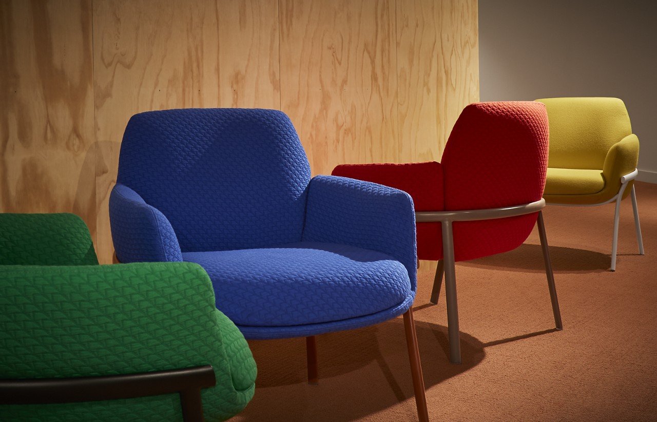 Four colorful modern chairs in green, blue, red, and yellow arranged in a room with wooden panel wall and carpeted floor.