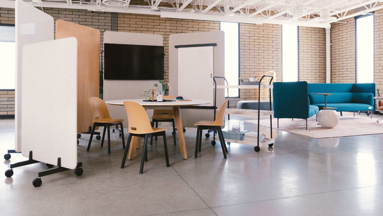 Modern office space with a round table, four chairs, a rolling cart, a lounge area with a blue sofa, and large windows.