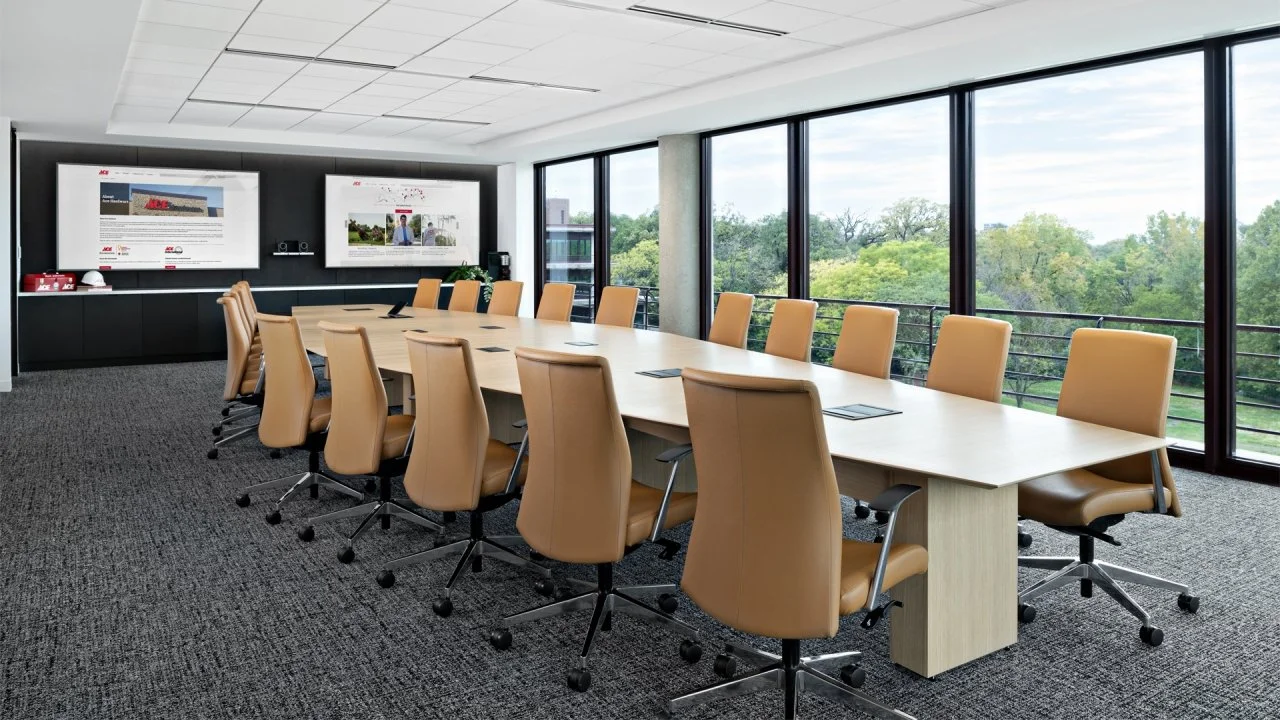 Modern conference room with a long wooden table surrounded by beige rolling office chairs, large windows showing trees outside, and two monitors on the wall.