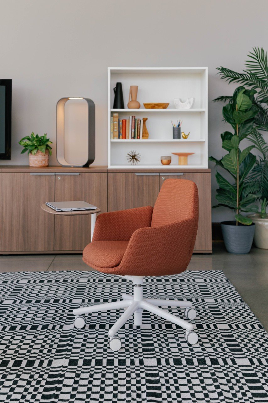 Modern home office with orange swivel chair on black and white patterned rug, wooden sideboard with decorative objects, white bookshelf with books and decor, and large potted green plants.