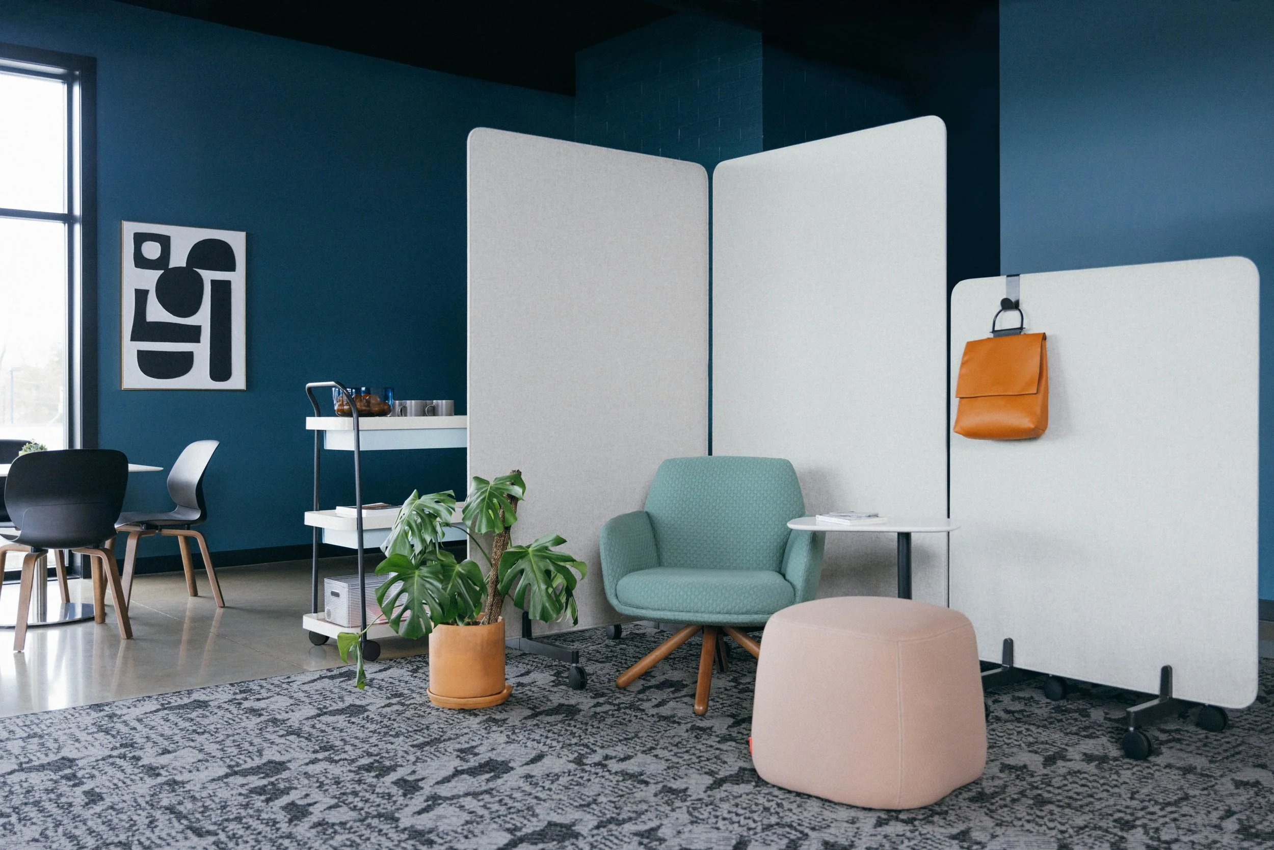 Modern office lounge with light green armchair, plant, white privacy screens, orange bag hanging on the screen, beige ottoman, and patterned rug, with navy blue walls and a black and white artwork.
