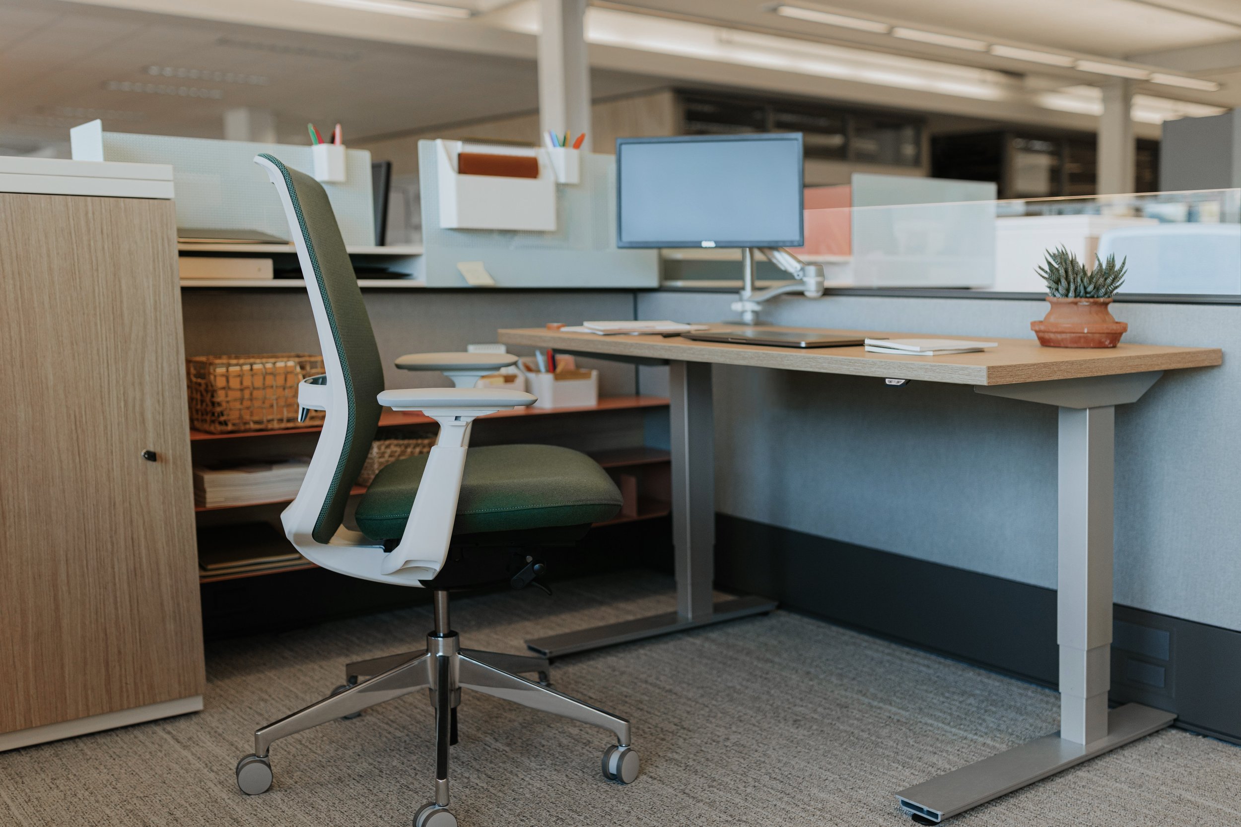 Empty office workspace with a green office chair, a desk with a small plant, computer monitor, and office supplies.
