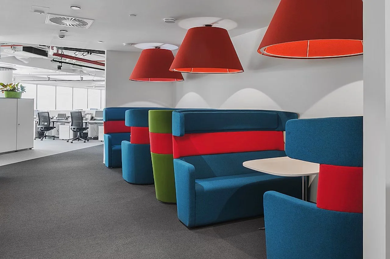 Office lounge area with blue and green booth seats featuring red bands, large red pendant lamps, carpeted floor, and open workspace with office chairs and desks in the background.