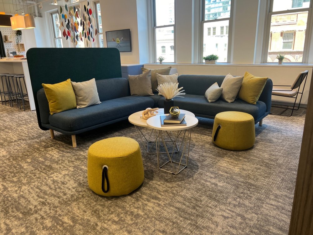 Living room with a dark blue sofa, yellow and beige cushions, two yellow ottomans, white round tables with decorative items, hanging art, large windows, and modern decor.